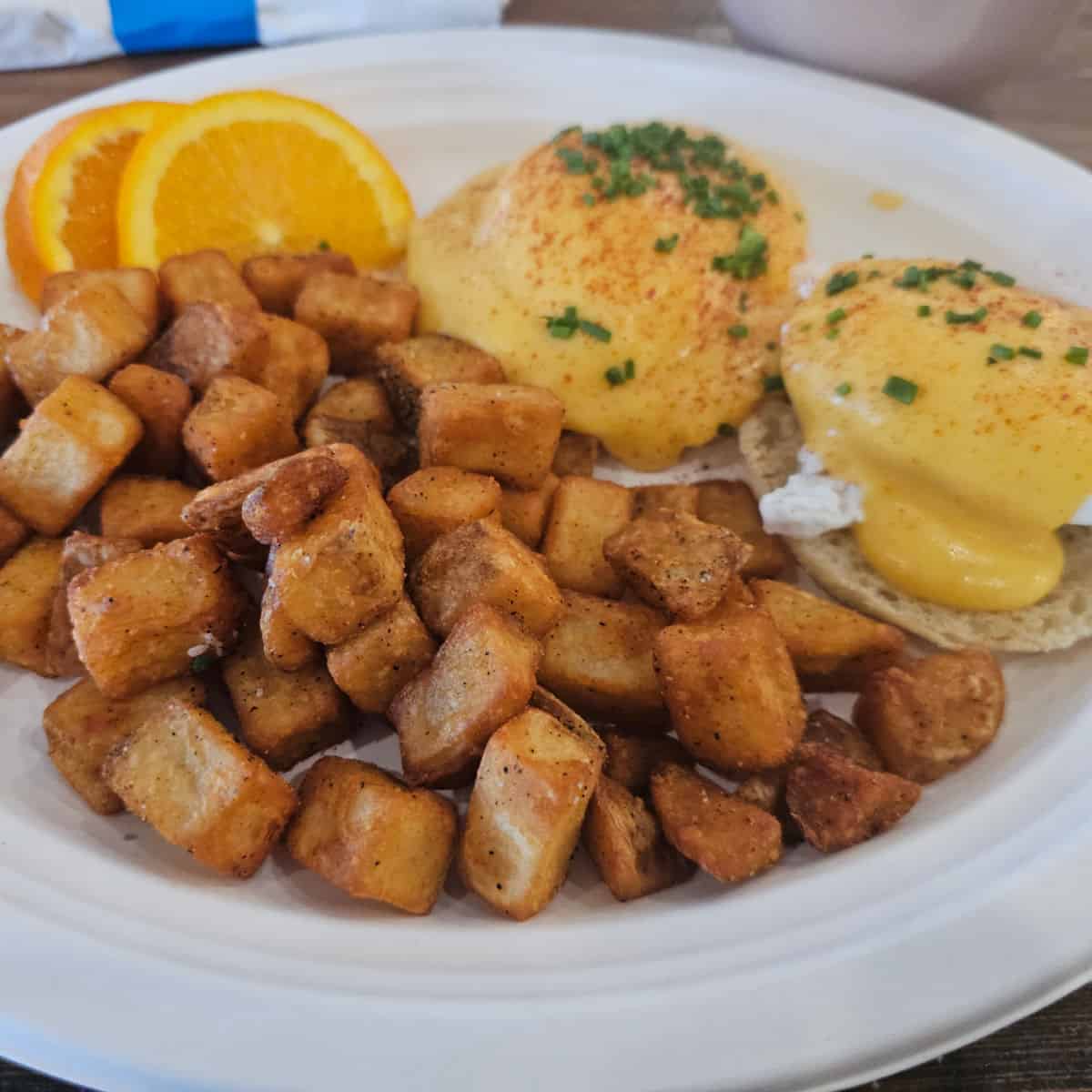 white plate with eggs benedict, home fries, and two orange wheels
