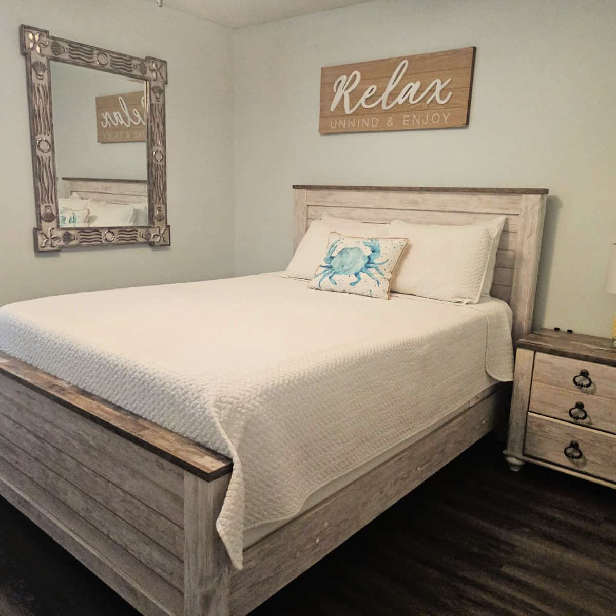 Bedroom with a bed with wooden frame, white linens, and a crab pillow, relax unwind and enjoy sign above the bed
