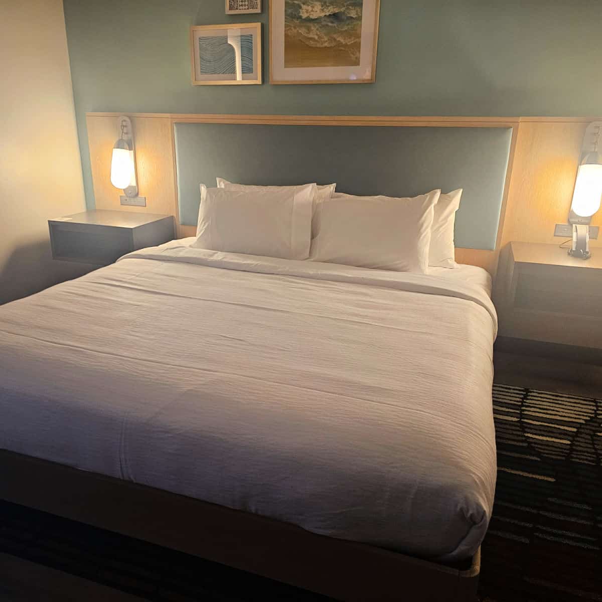 king bed with white linens, two lamps on nightstands, a couple pieces of beach art above the bed