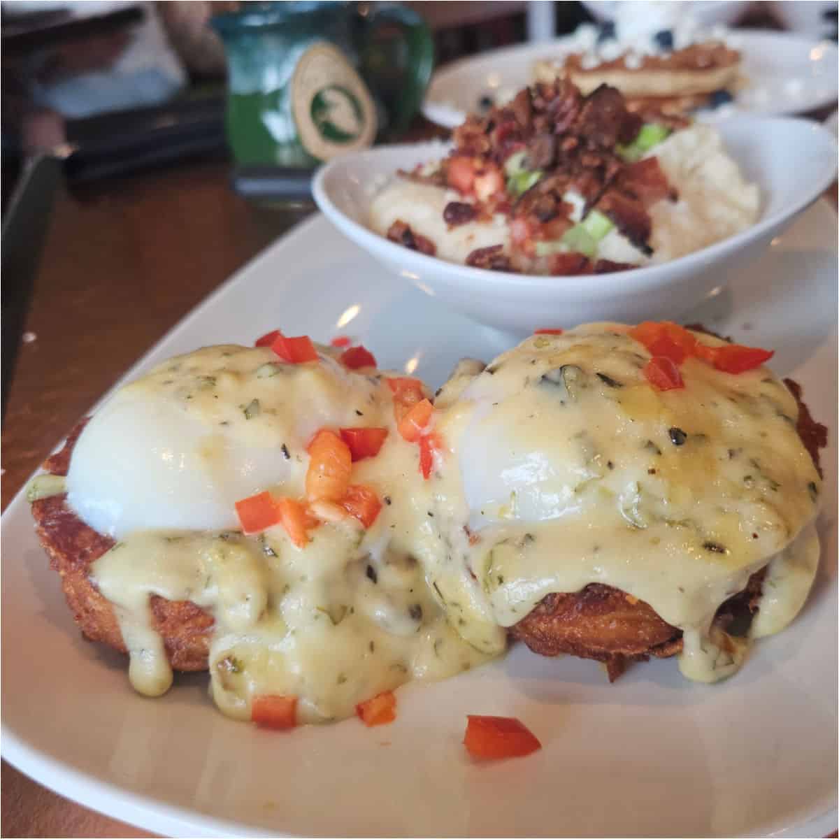 Hashbrown benedict covered in hollaindaise sauce on a white platter with a bowl of loaded grits