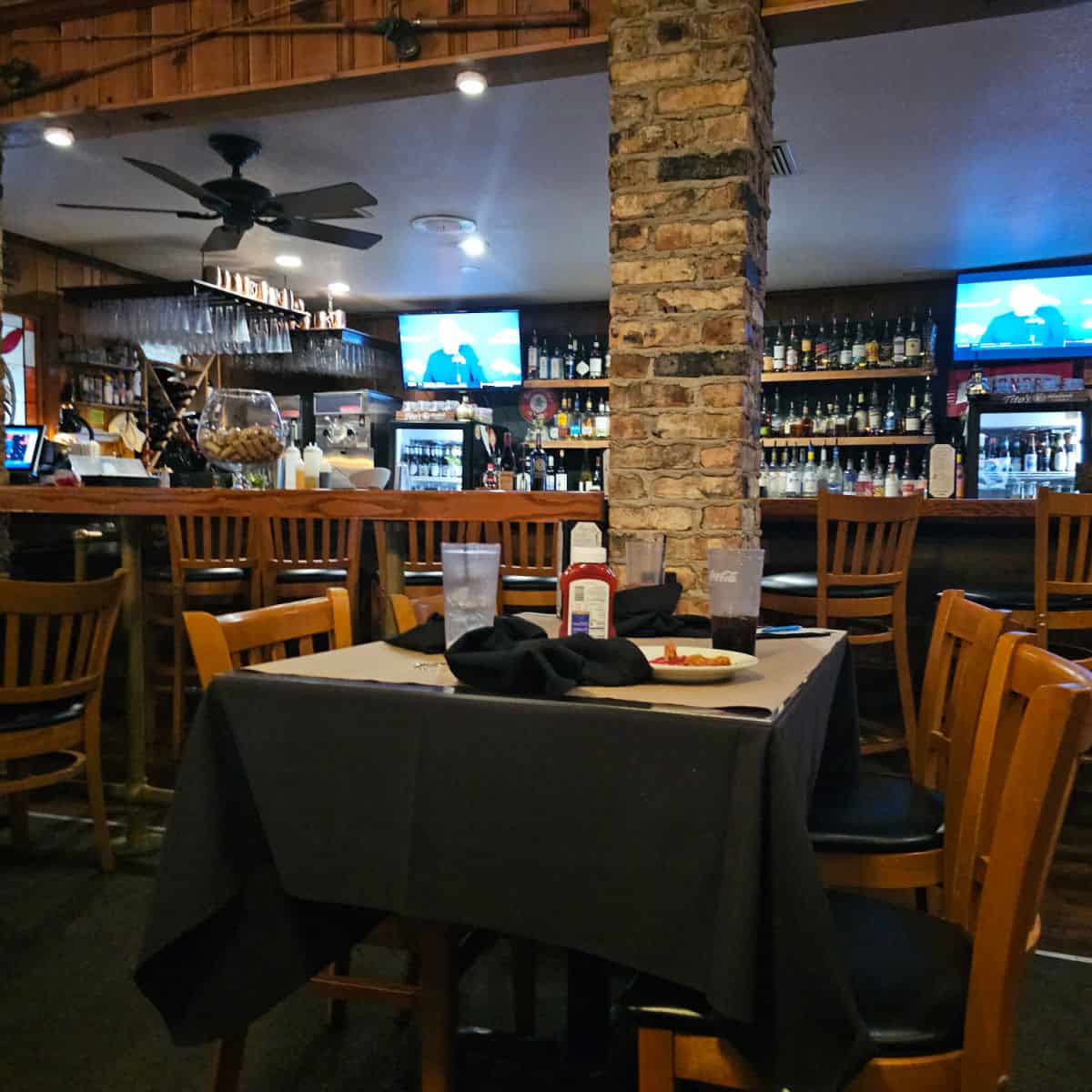 indoor bar area wtih a wall of bottles between tv's, tables and chairs next to a brick column