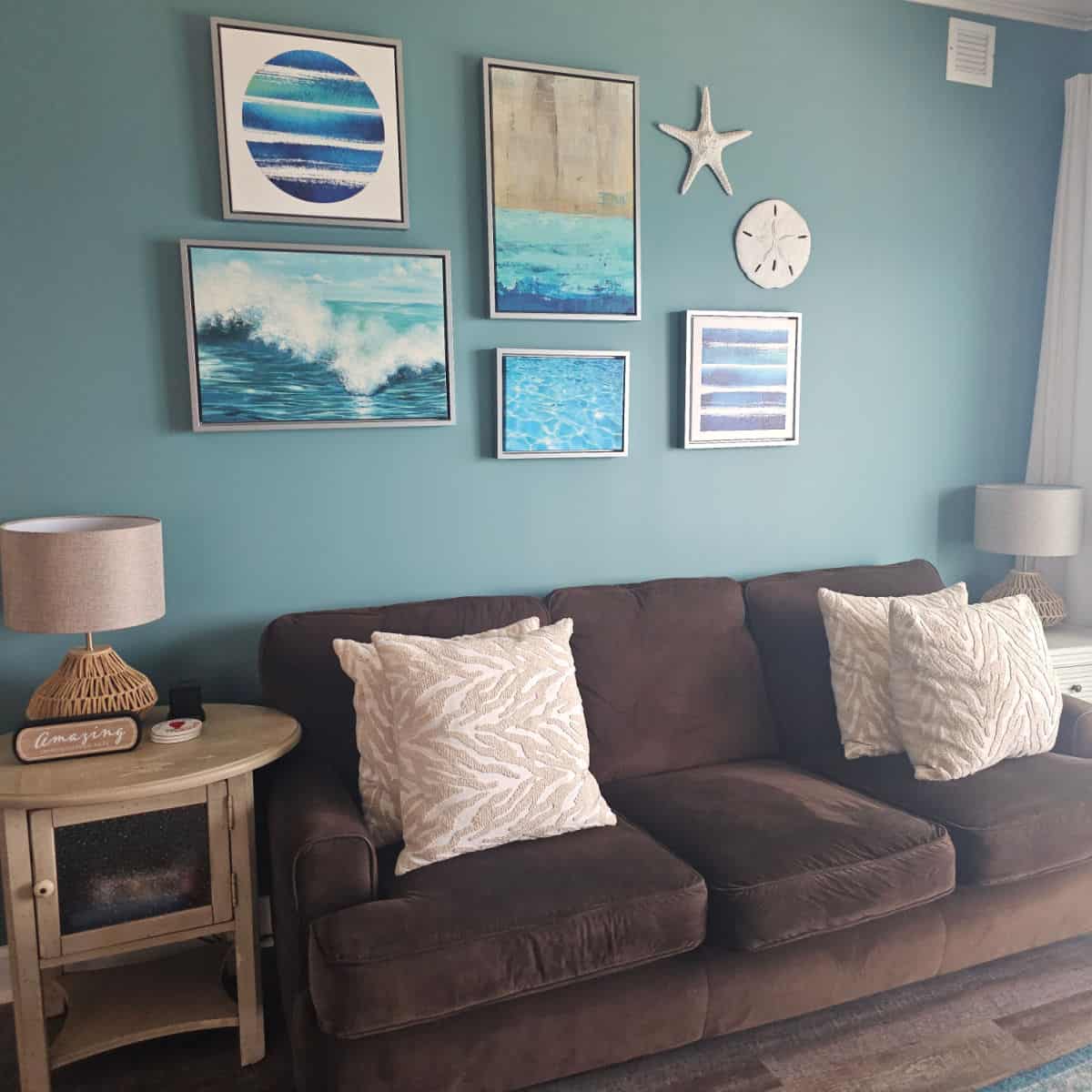 living room area with coastal beach art on the blue wall behind a brown couch with pillows, wooden table with lamp
