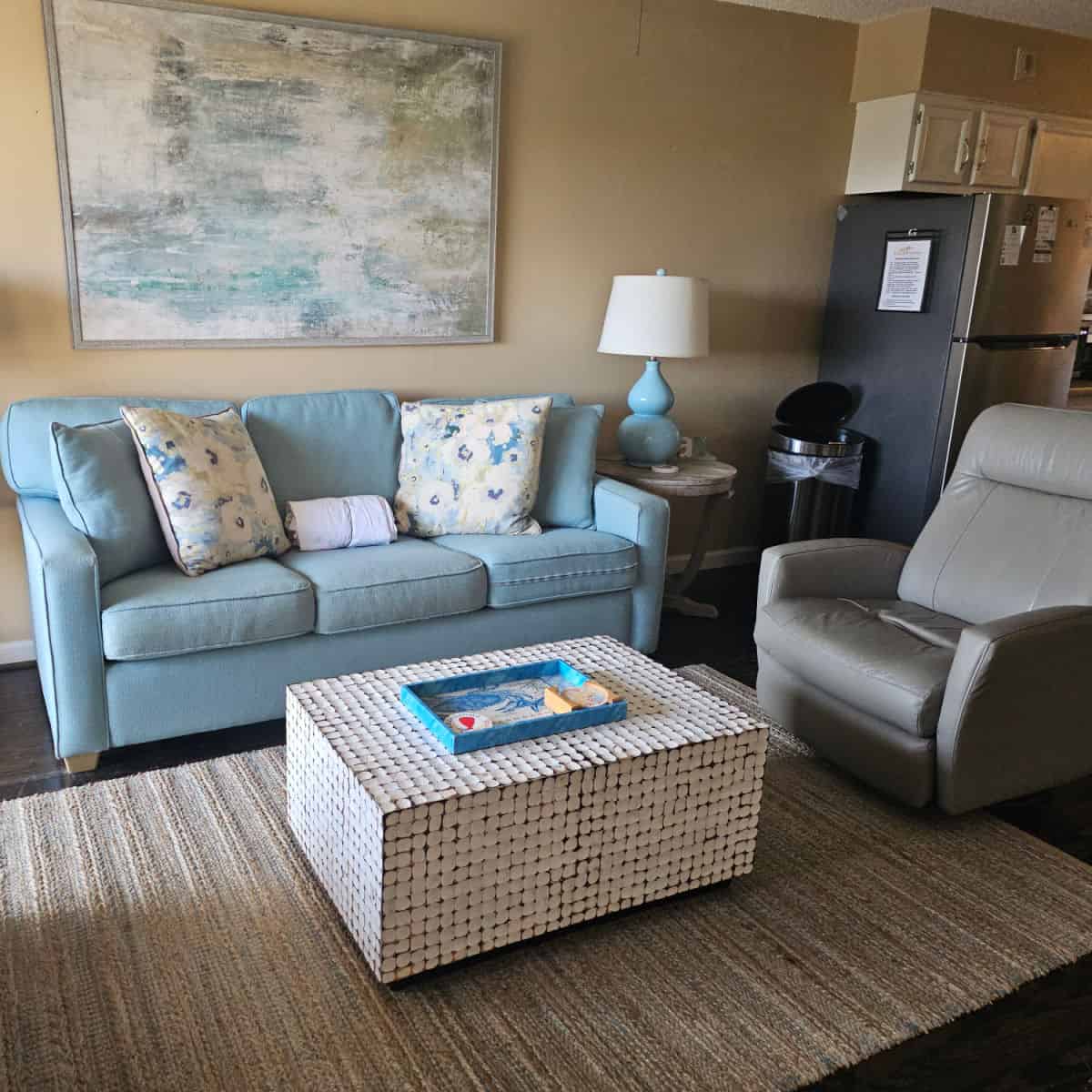 Living room with blue couch, white table with a large tray, grey leather chair and art over the wall. Edge of kitchen in the right corner with a stainless steel fridge