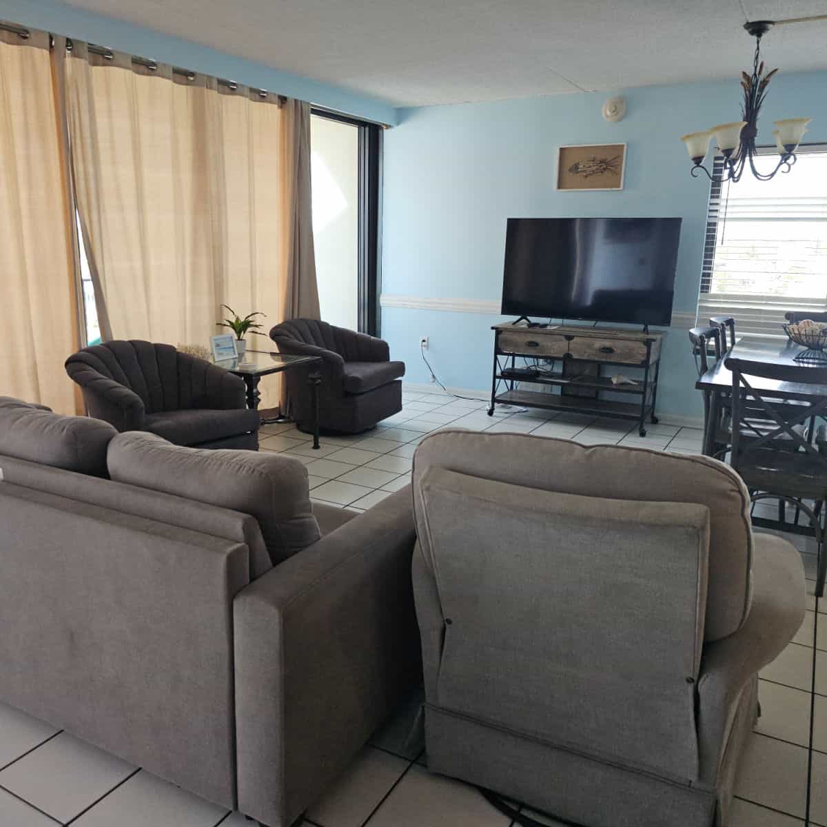 Living room with couch and a couple of chairs, tv on a wooden stand, dining room table with chairs under a lamp, shades over the windows