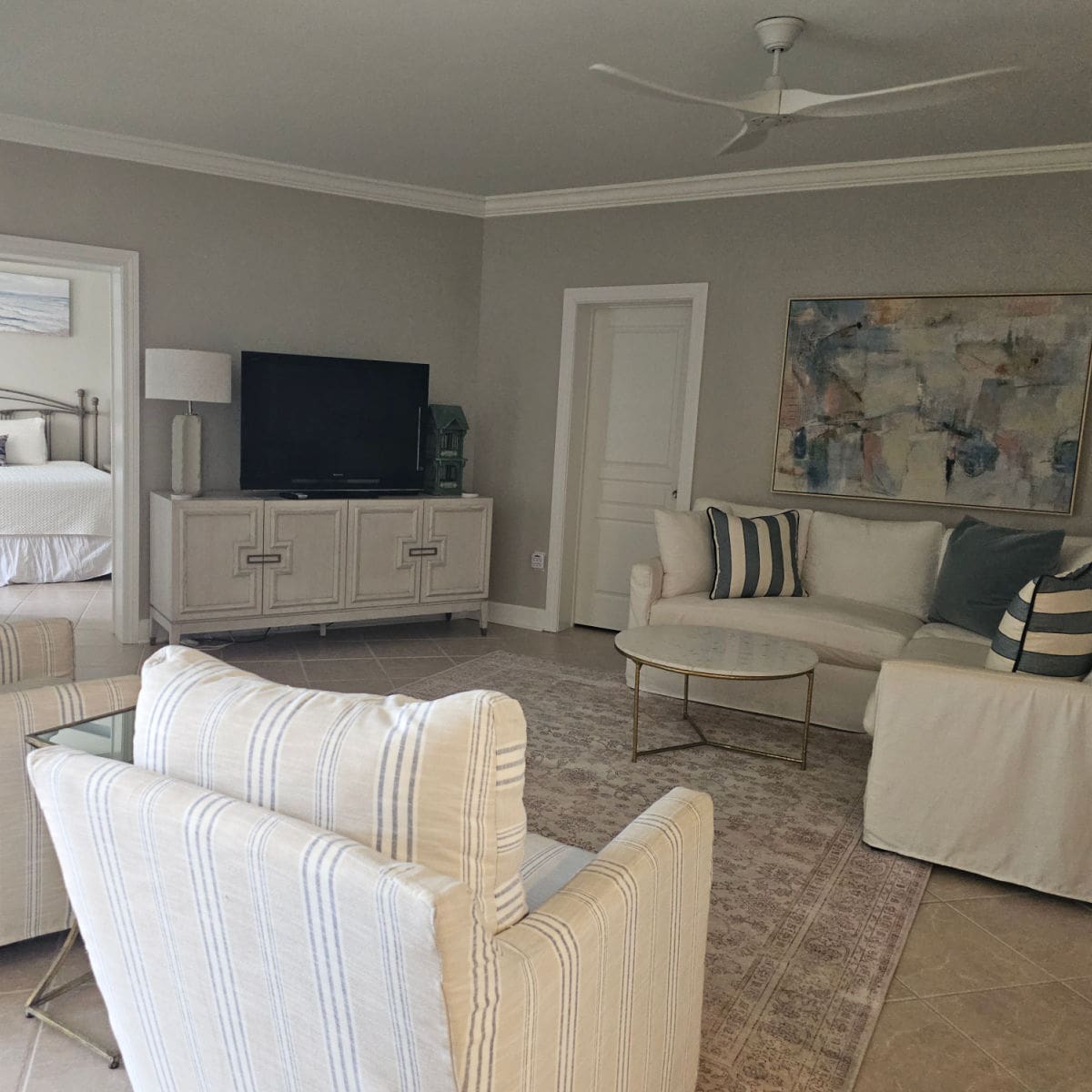 living room with white couch with blue and white pillows, round table by the couch, large art piece above the couch, flatscreen tv on a cabinet near the wall, a couple of striped chairs opposite the couch