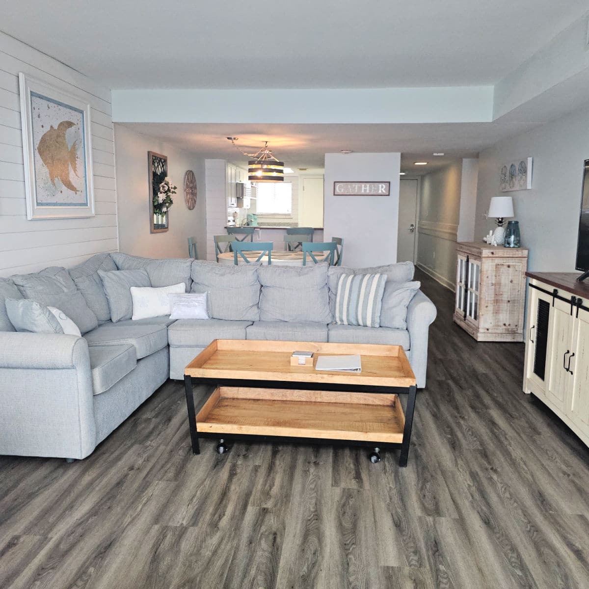 Condo with a grey wrap around couch and table, looking past the couch to a dining room table and kitchen area, hall running down the side of the room