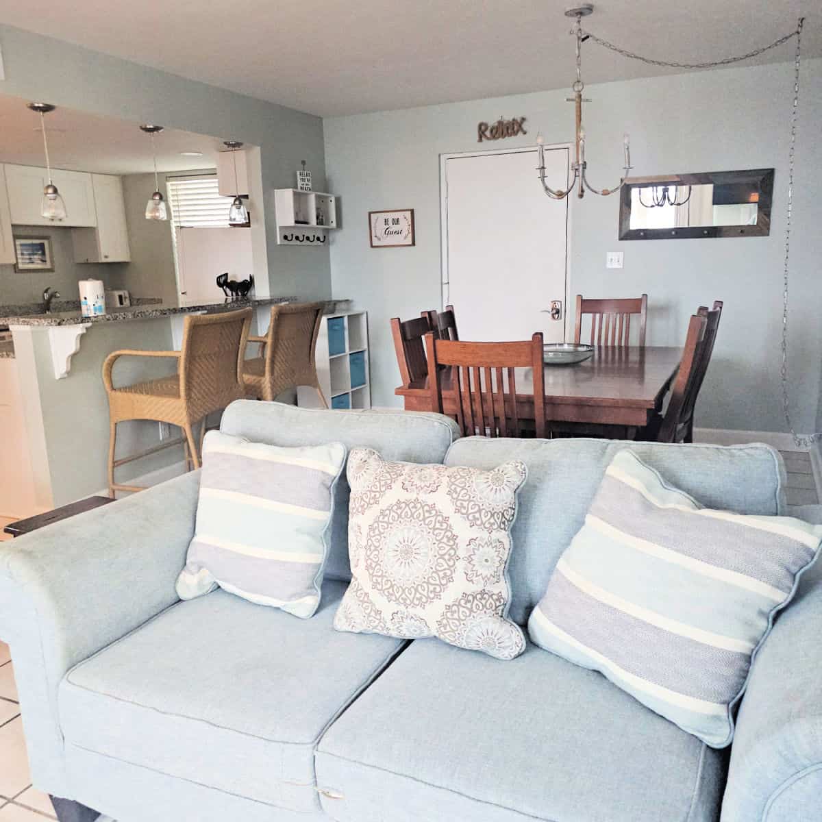grey couch with pillows, dining room table behind it with a chandelier, Kitchen on the left side with tall bar chairs