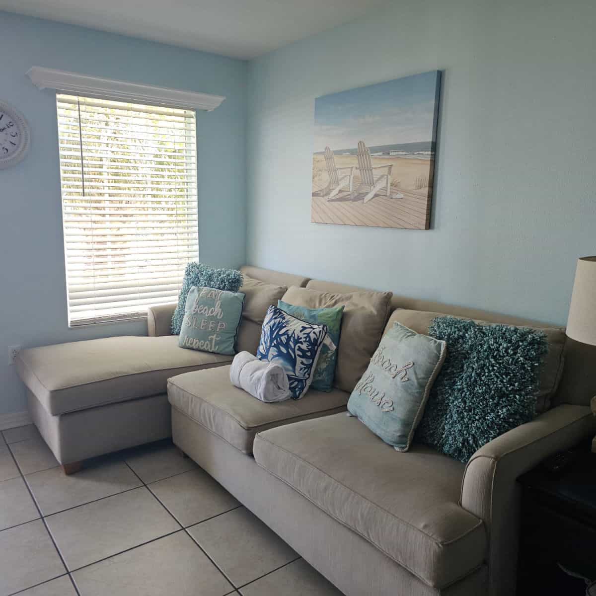 Taupe couch with pillows next to a table with a lamp, picture on the blue wall with adirondack chairs