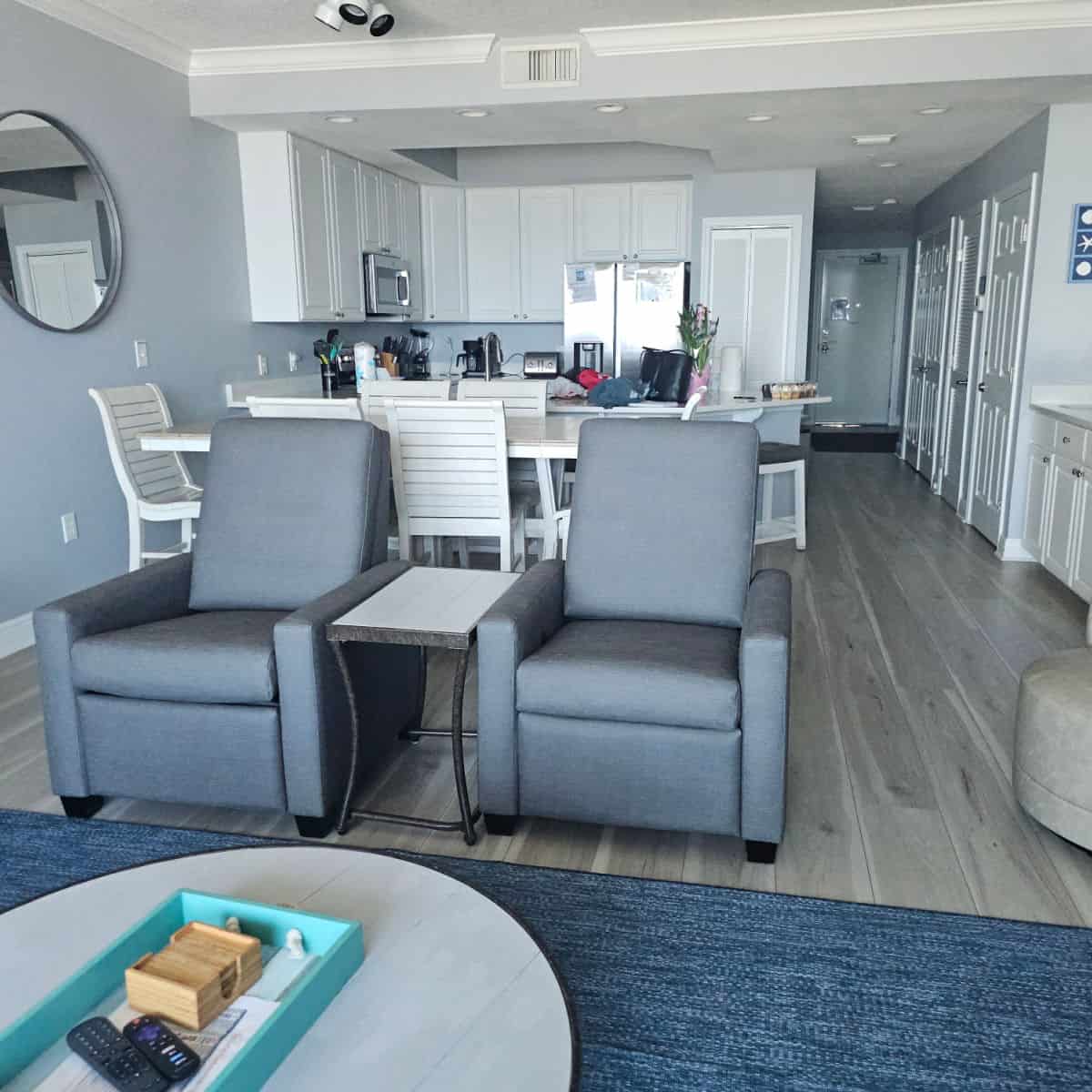 condo unit with two chairs and a table at the front, table with white chairs, then kitchen, hallway on the right side leading to the front door