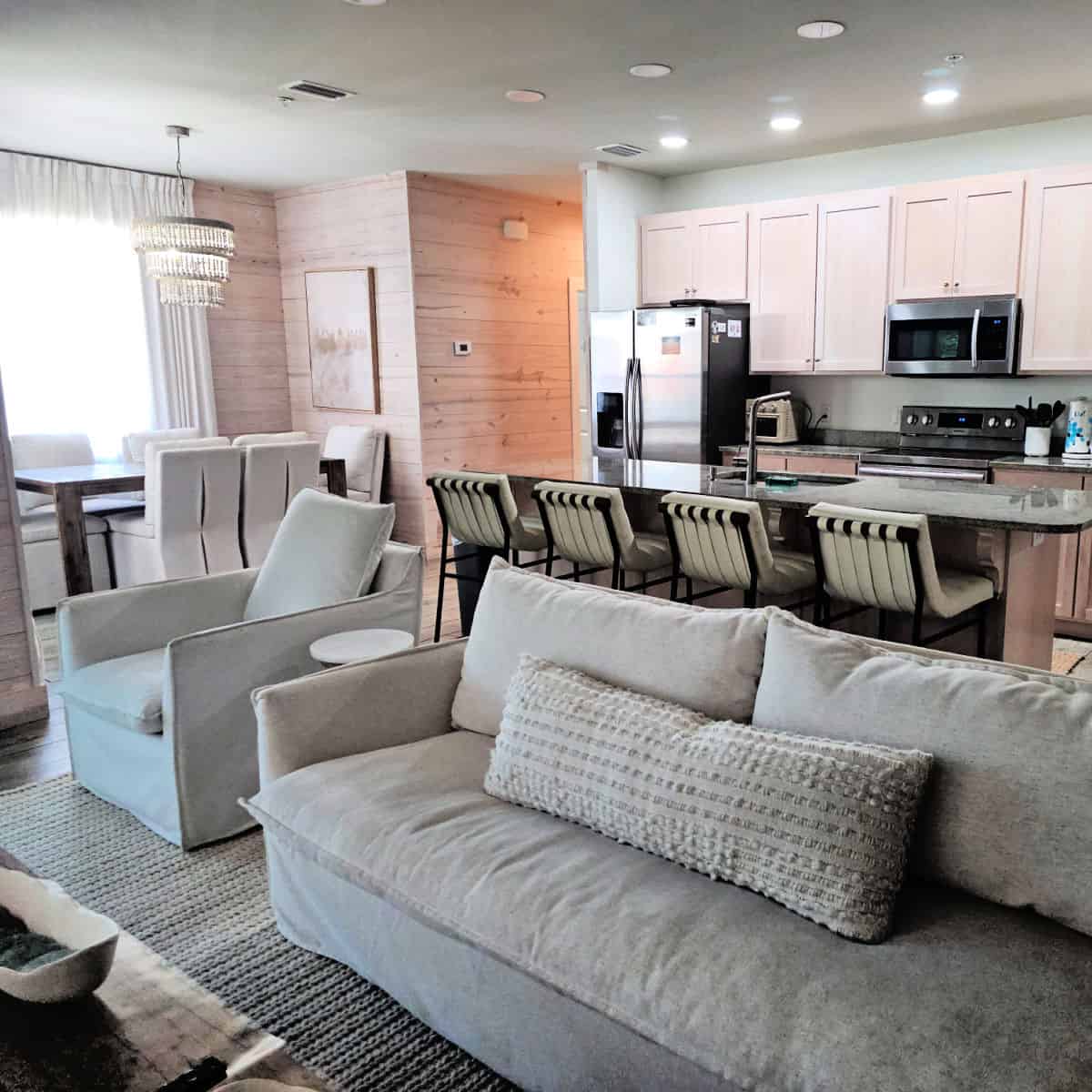 looking over the living room couches with fluffy pillow towards the kitchen with bar chairs and lightly tanned cabinets, dining room table on the left side with a chandelier