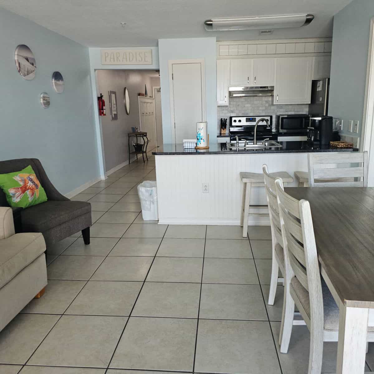 Dining room table on the right side of the image, kitchen with stainless steel appliances in the background, hall with paradise sign above it, and then a couch and chair on the left