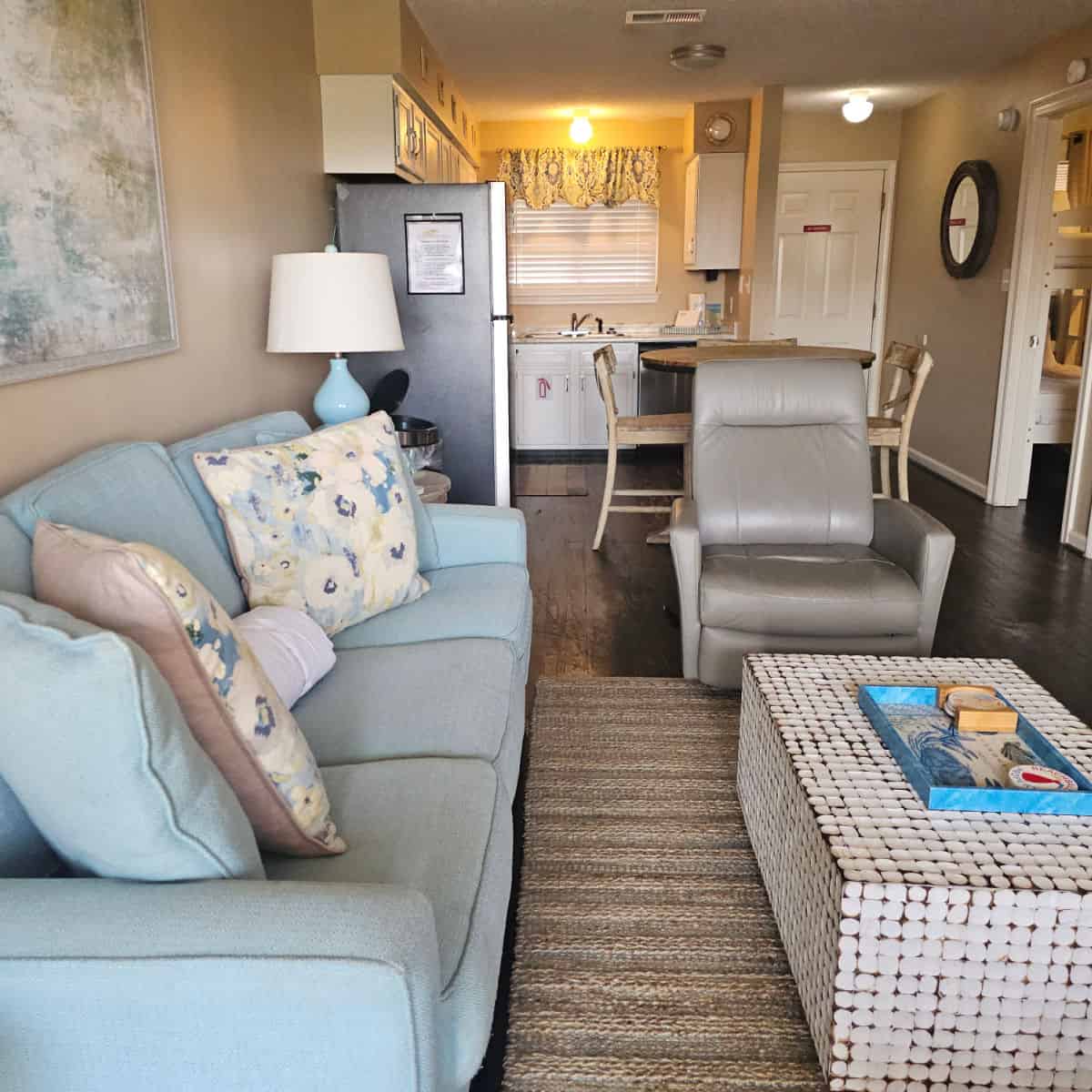 living room with blue couch and pillows, white square table, leather chair, kitchen area with table and chairs and cabinets leading towards the door