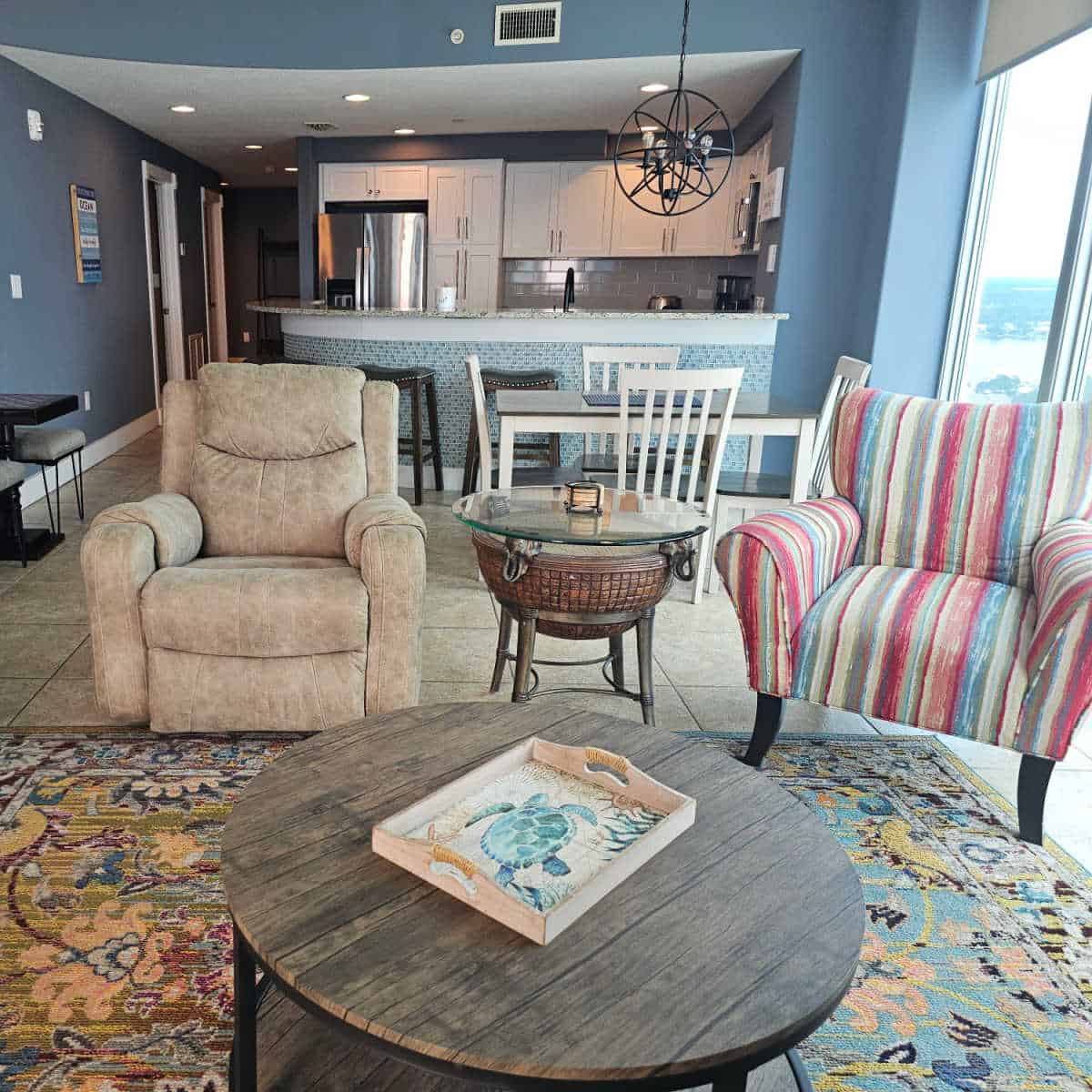 wooden table with a turtle art tray, two comfy chairs with a glass table in between them. Looking back towards a dining room table with white chairs and a kitchen with white cabinets, windows along the right side