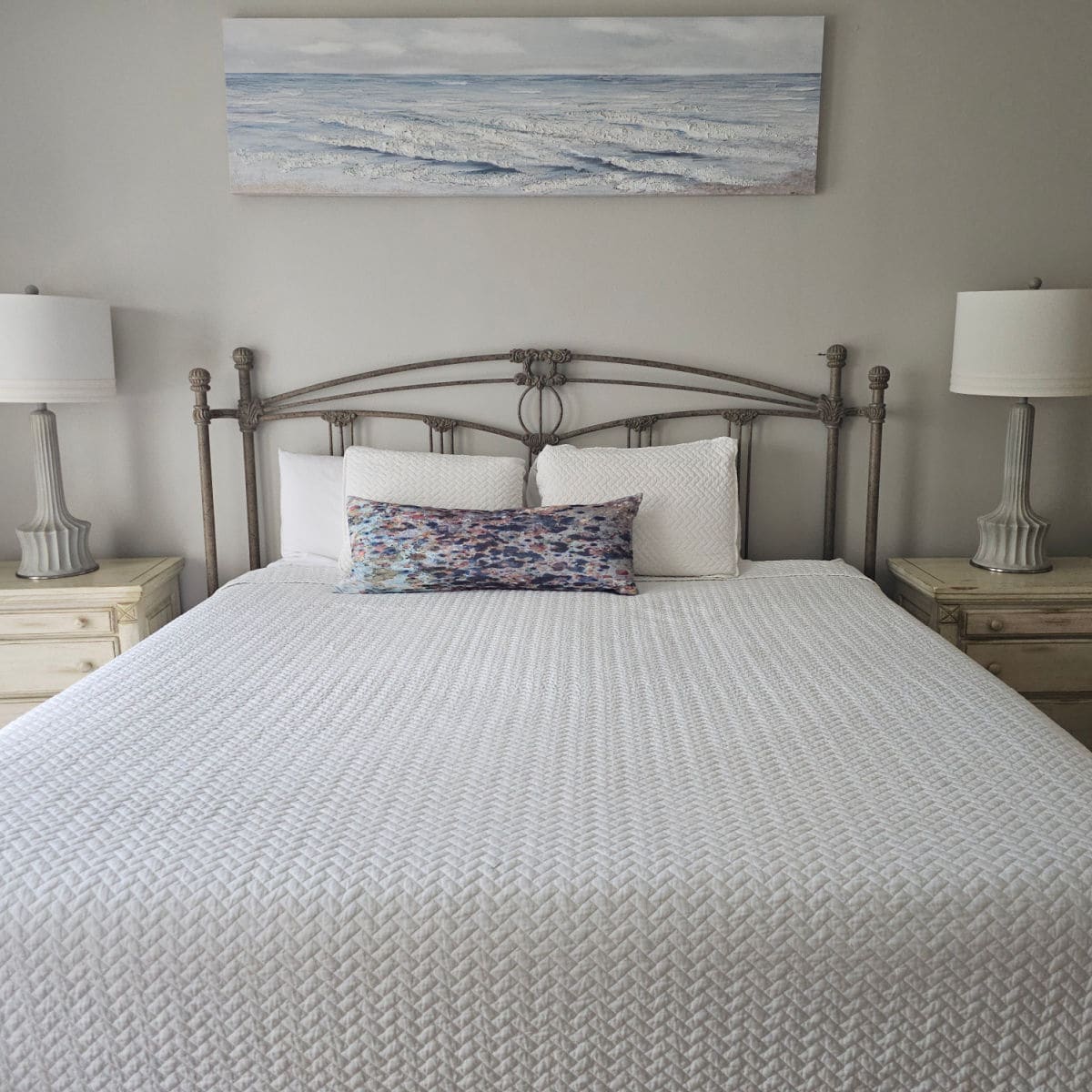 large bed with white cover and a couple of pillows, scenic beach art above the bed, 2 night stands with lamps next to the bed