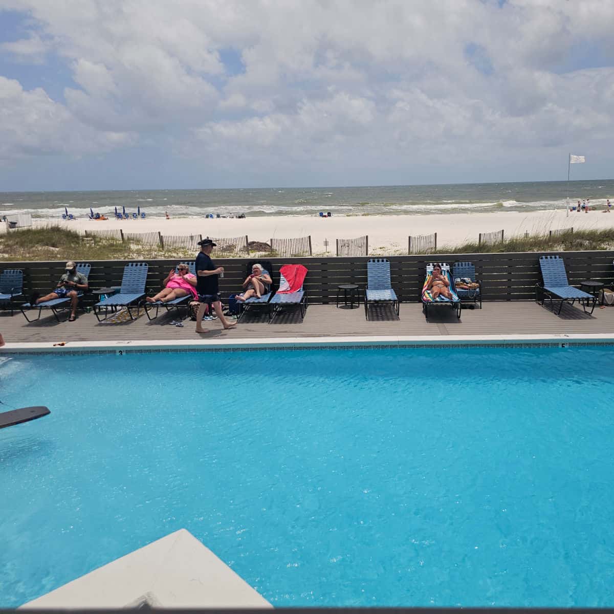 outdoor pool with chairs lining the pool deck, people sitting in the chairs, Gulf waters and beach behind the pool deck