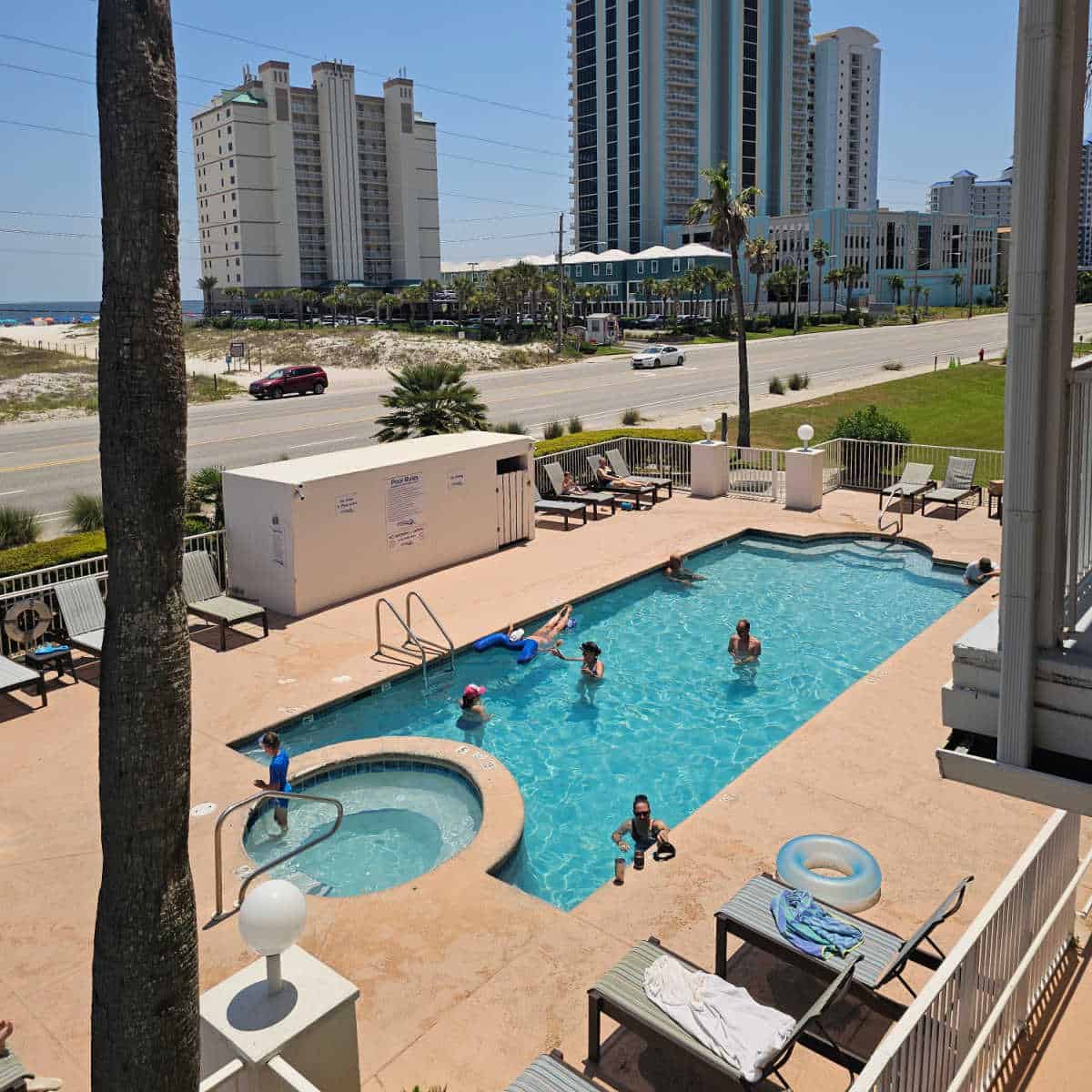 outdoor pool with people in the water, chairs around the pool deck, with a fence around the pool. road running along the length of the picture with condo buildings on the backside and the Gulf waters in the distance