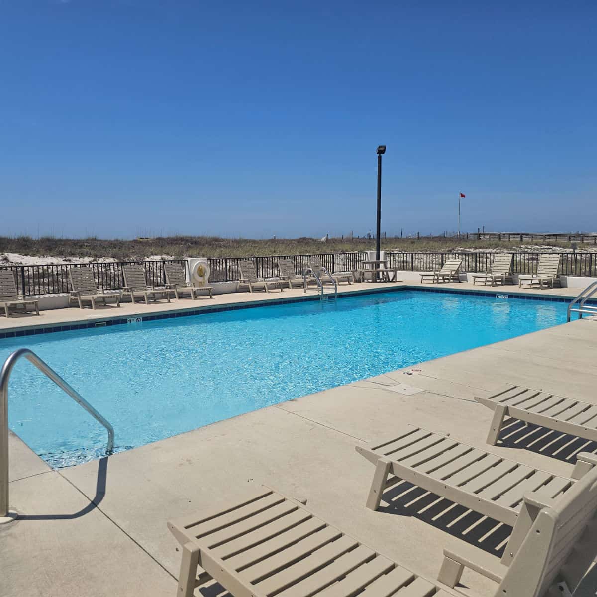 Outdoor pool with lounge chairs around the pool deck, sand dunes behind the fence and flag flying in the distance