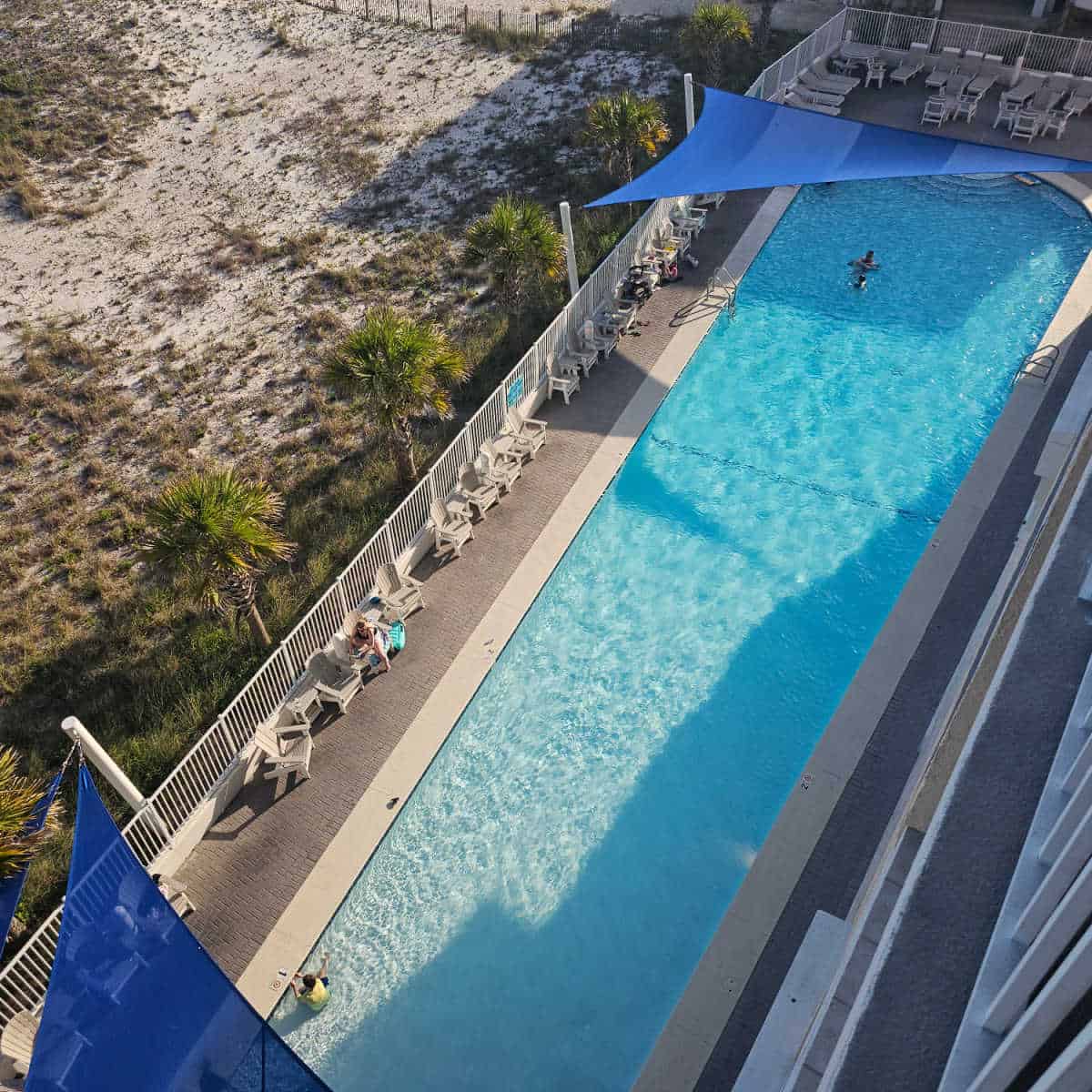looking over an outdoor pool with a couple people in the water, chairs along the pool deck, sand dunes on the side of the pool