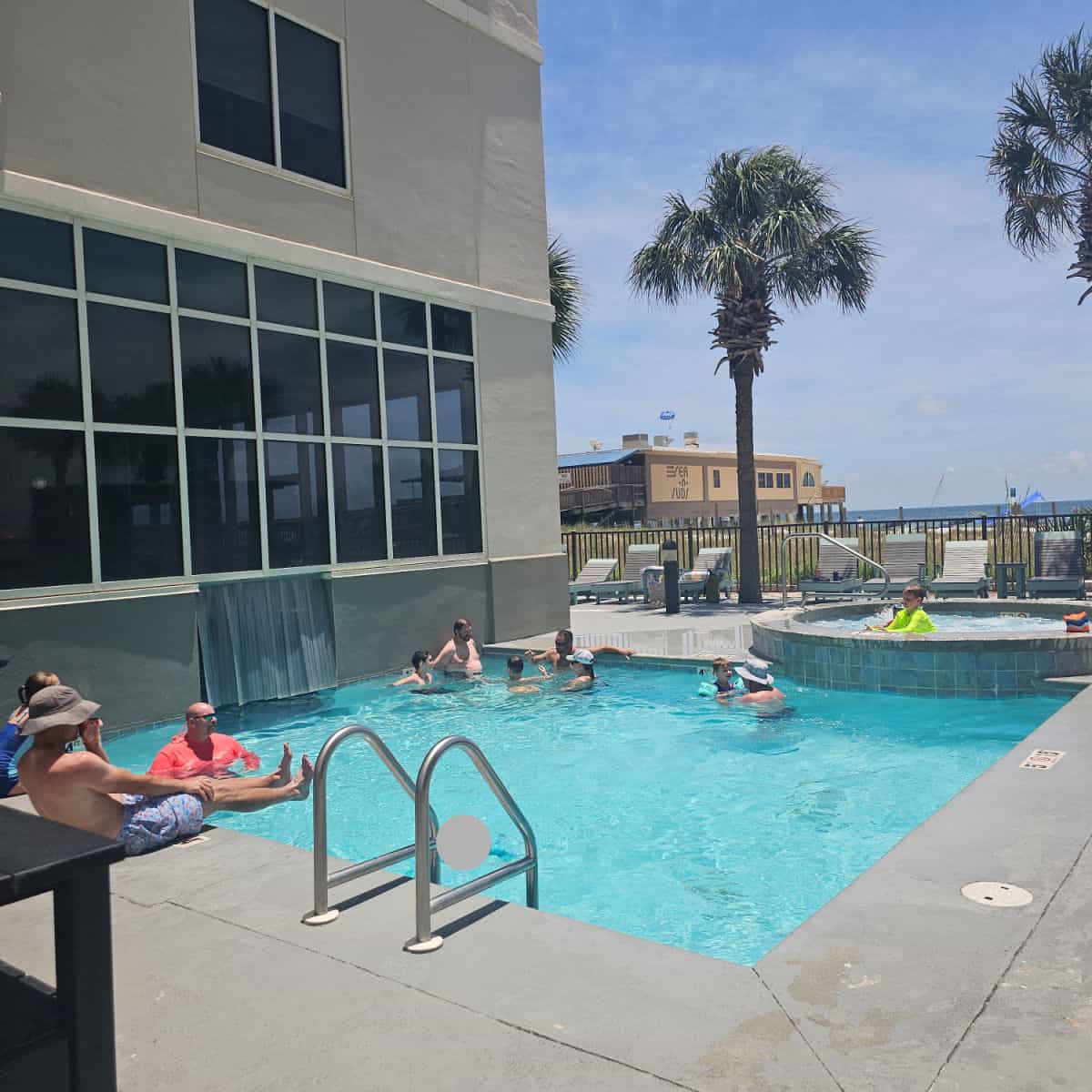 square outdoor pool with people in the water, connected to a circle hot tub, on a pool deck with lounge chairs, Sea n Suds restaurant in the background
