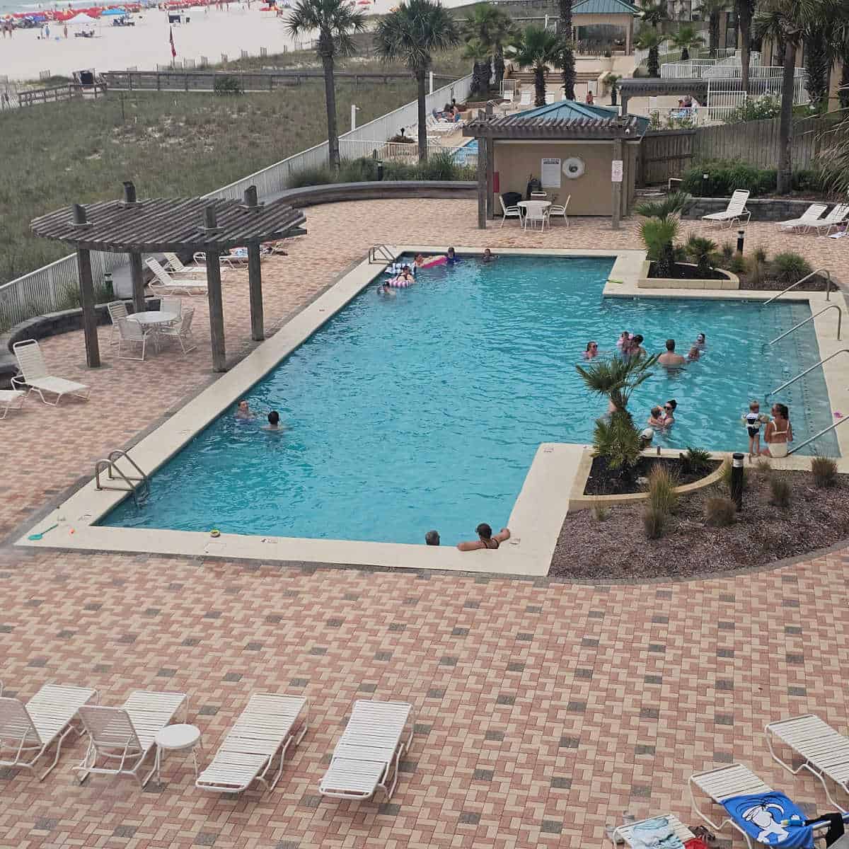 outdoor swimming pool with people in it, lounge chairs on the pool deck, beach in the top left corner