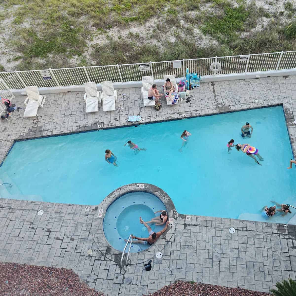 Looking down on an outdoor pool with people playing in the water, hot tub and deck chairs