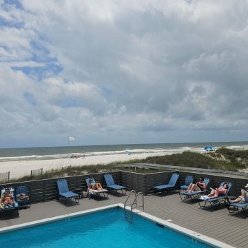 Corner of an outoor pool with people in blue lounge chairs around the pool deck looking out to the beach and Gulf waters on a cloudy day