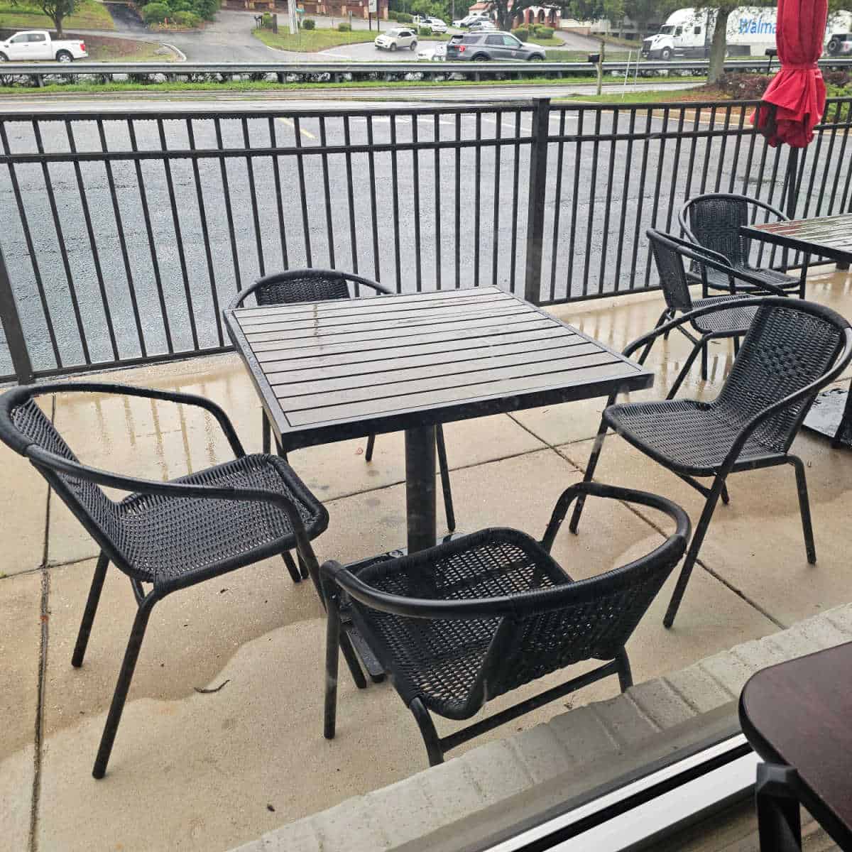 table and chairs outside on a patio with a black fence then parking lot and road in the background