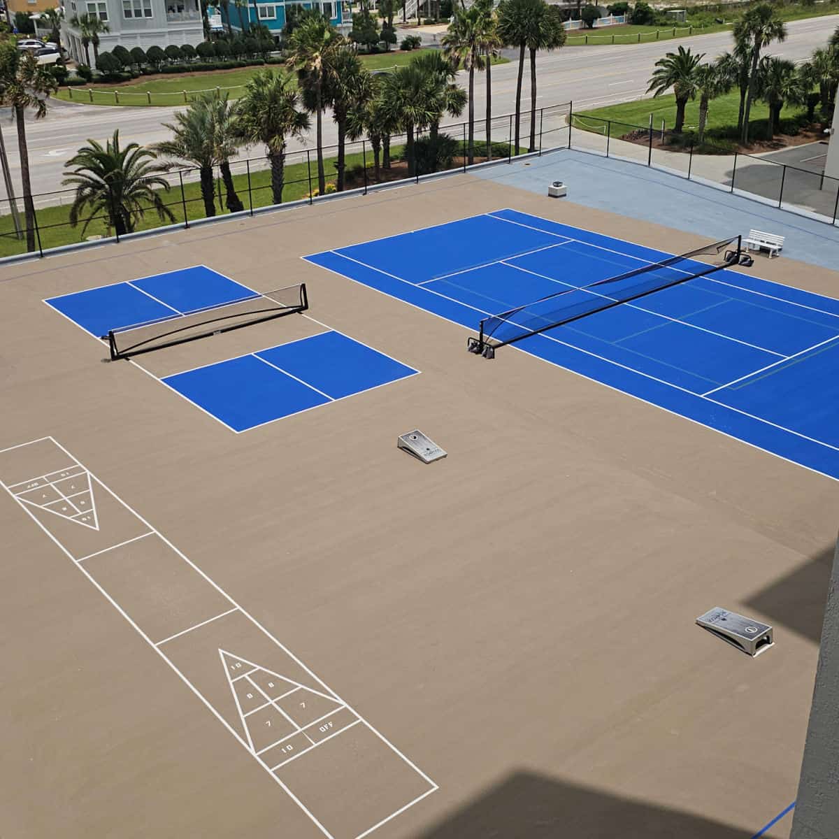 looking down over an amentieis deck with pickleball court, tennis court, and shuffleboard. palm trees outside of the fence lining the courts