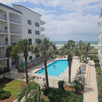 View looking over the outdoor pool with palm trees around it, building on one side, and Gulf waters in the background