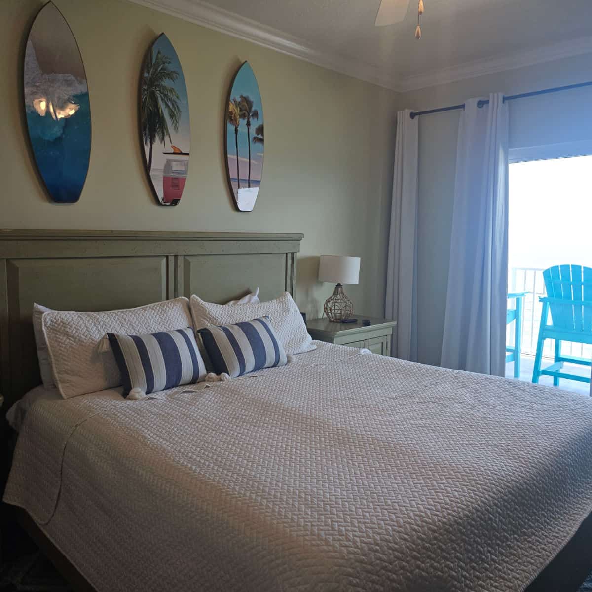 bedroom with white linen covered bed, three surfboards above the bed, window with adirondack chairs on the balcony