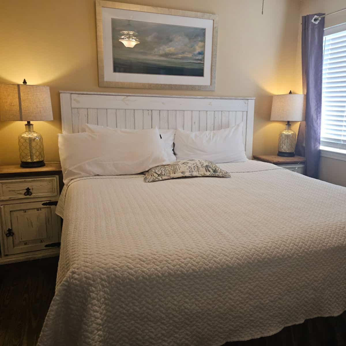 Bedroom with white linens and pillows, wooden nightstands with lamps and art above the bed