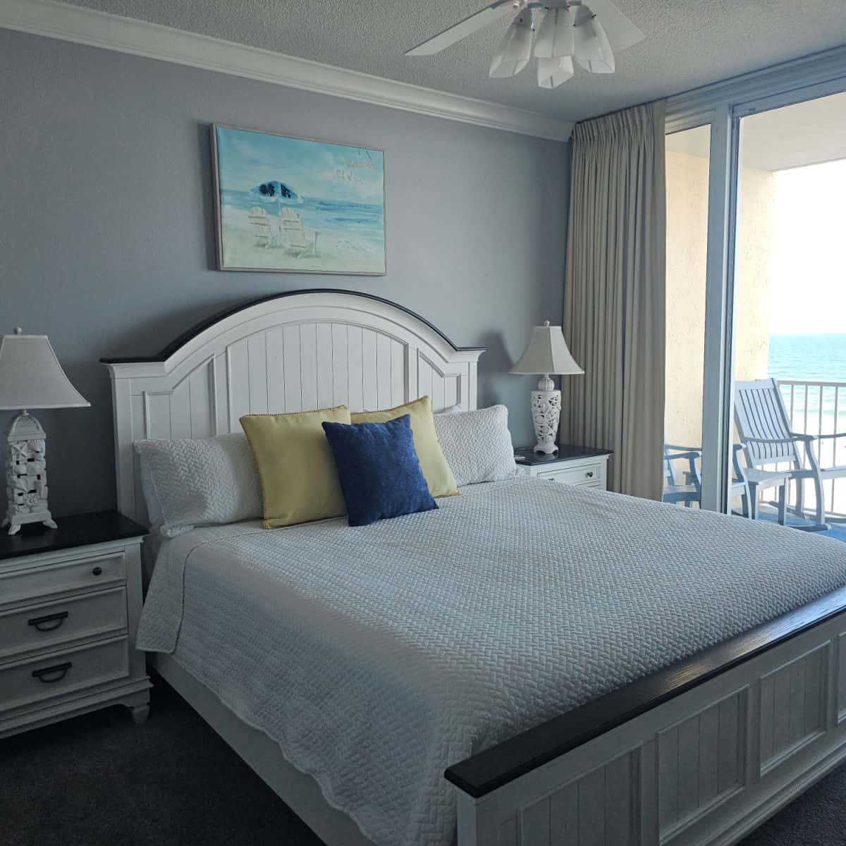 bedroom with large bed with white linens, nightstands with lamps, balcony with adirondack chairs in the background