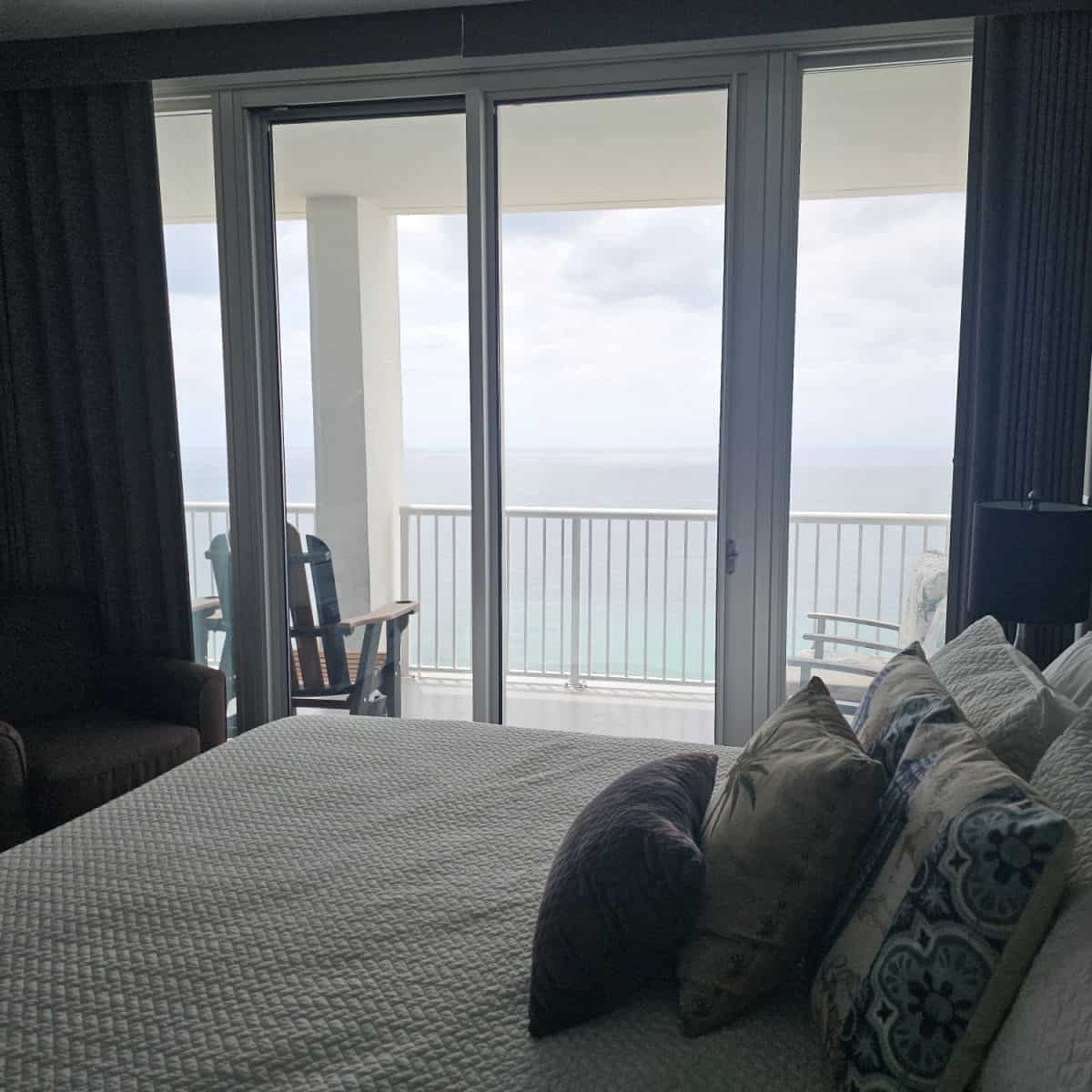 bed with lots of pillows and white linens with a glass door leading out to the balcony with views of the Gulf and an adirondack chair