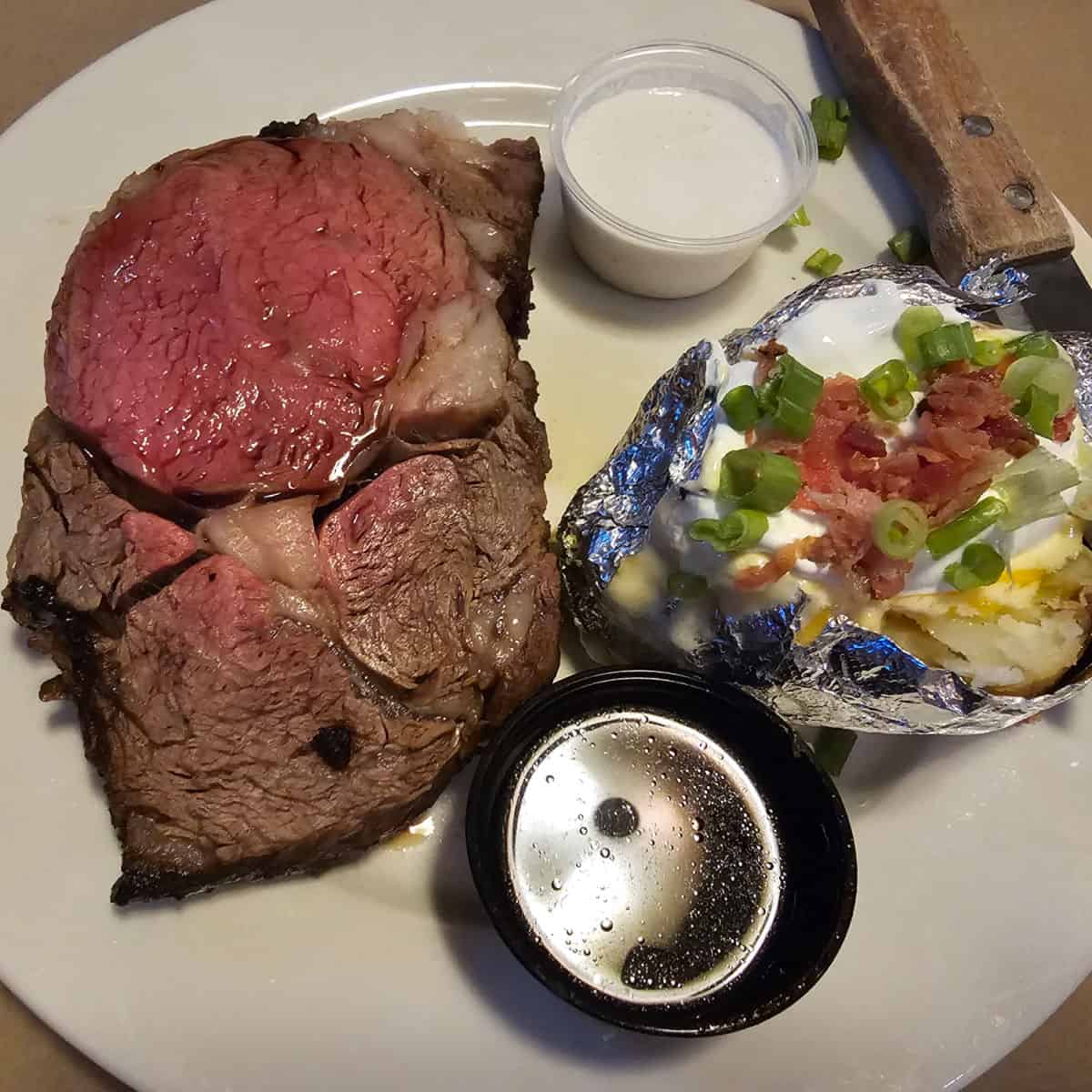white plate with a slice of prime rib, baked potato, au jus sauce, and knife