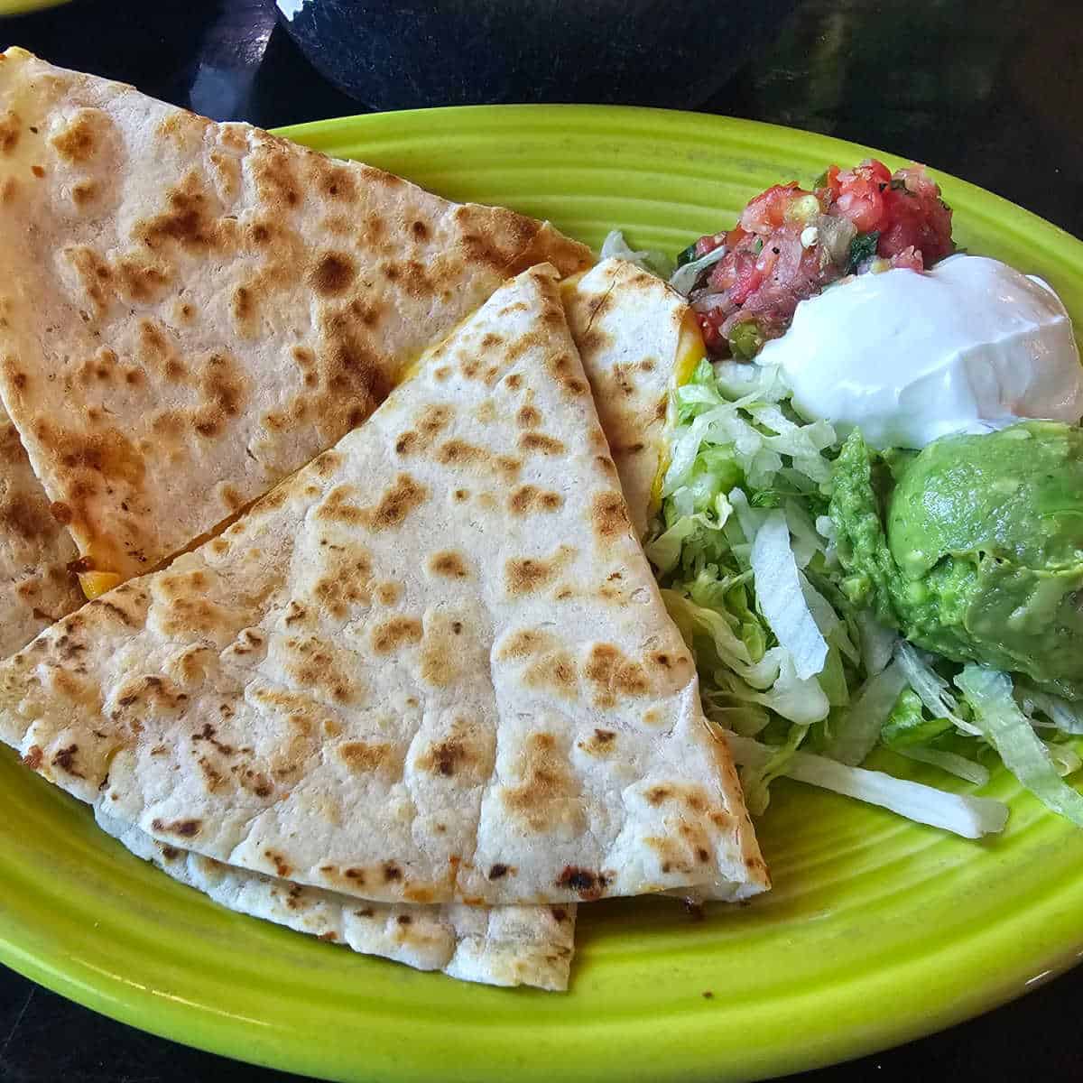 cheese quesadilla on a green plate with salsa, sour cream, and guac