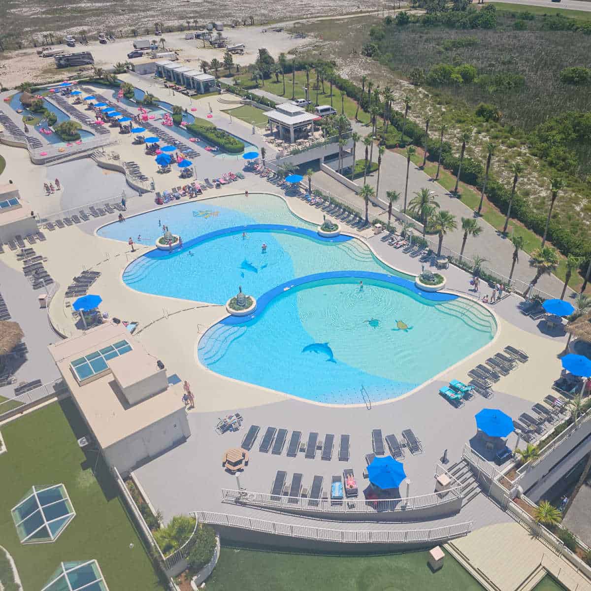 looking down over a large ool deck with three connected pools, lazy river, and cabanas, greenery and palm trees around the edge