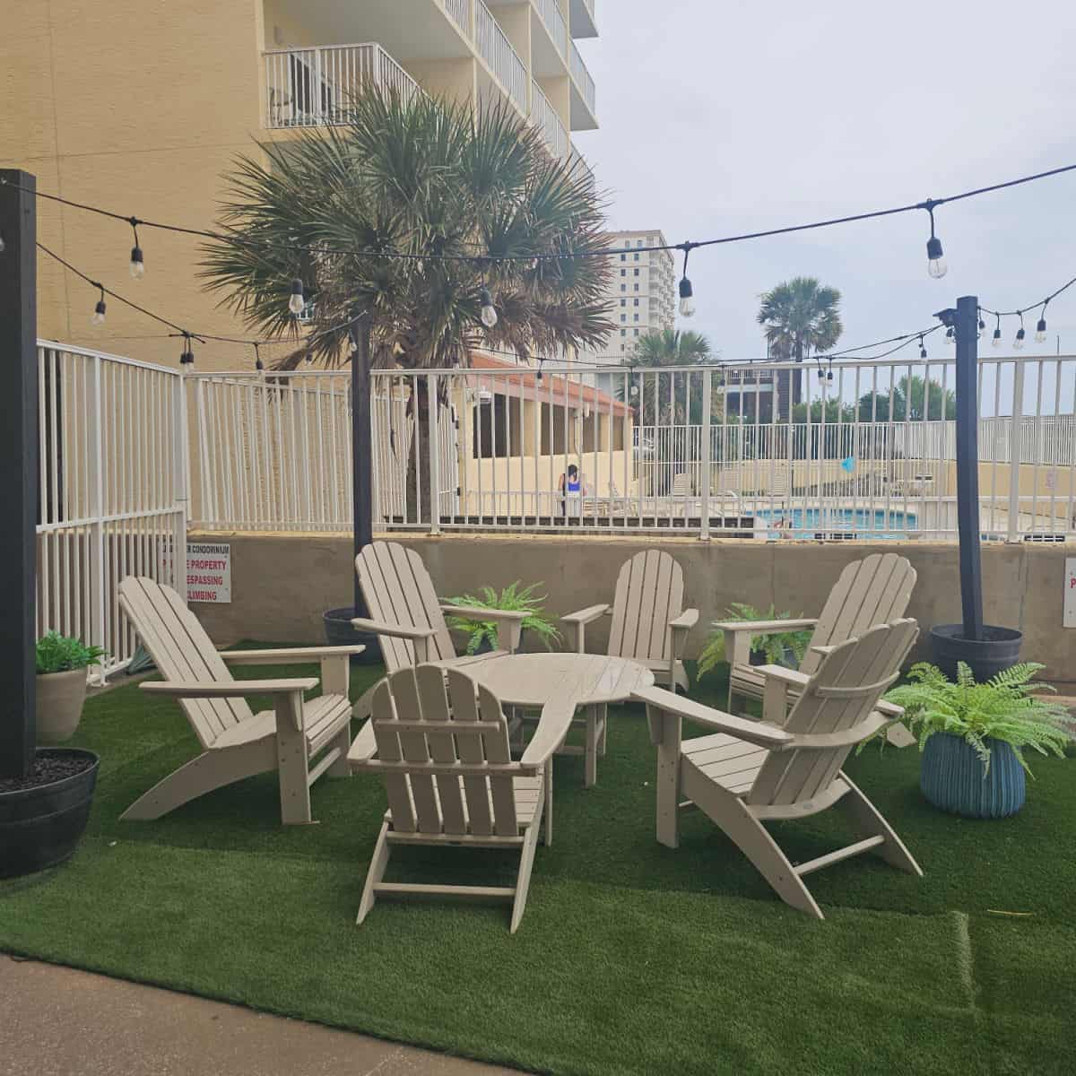 Adirondack chairs in a circle around a table with plants around them. Lights hanging from poles over the chairs