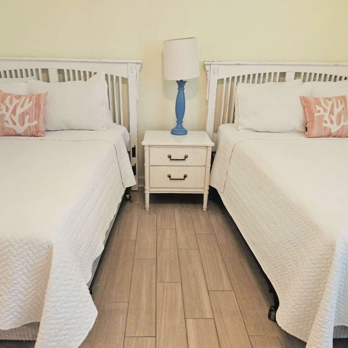 two beds with white linens and a pink coral pillow, nightstand between the bed with a lamp