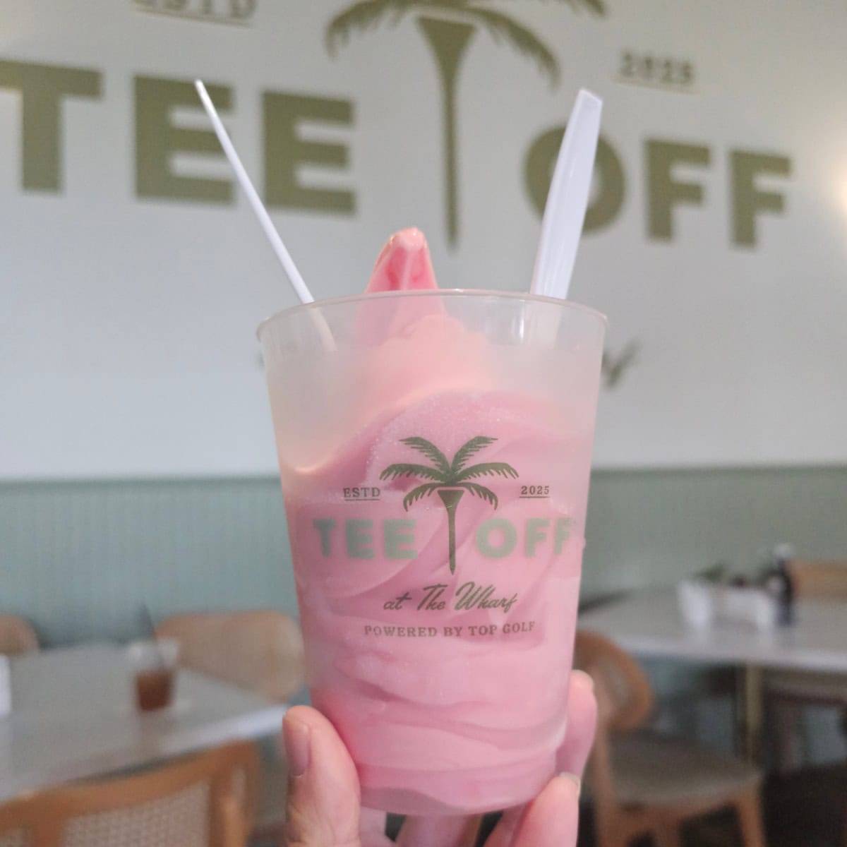 hand holding a Tee off at The Wharf glasss with a strawberry soft serve margarita in it. tables in the background