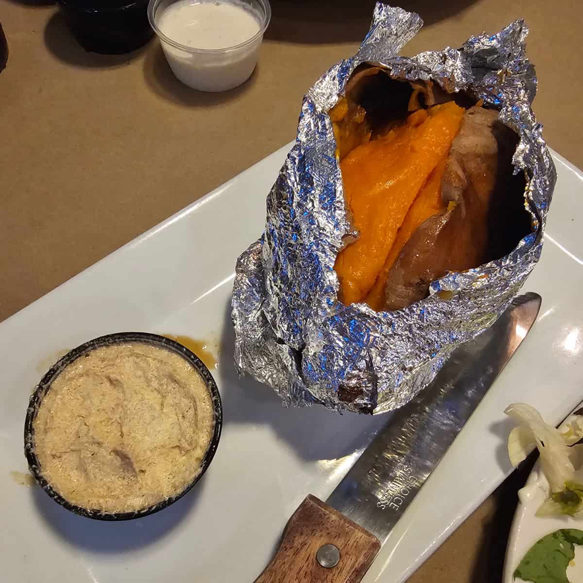 Sweet potato wrapped in foil on a white platter next to a knife and a container of honey cinnamon butter
