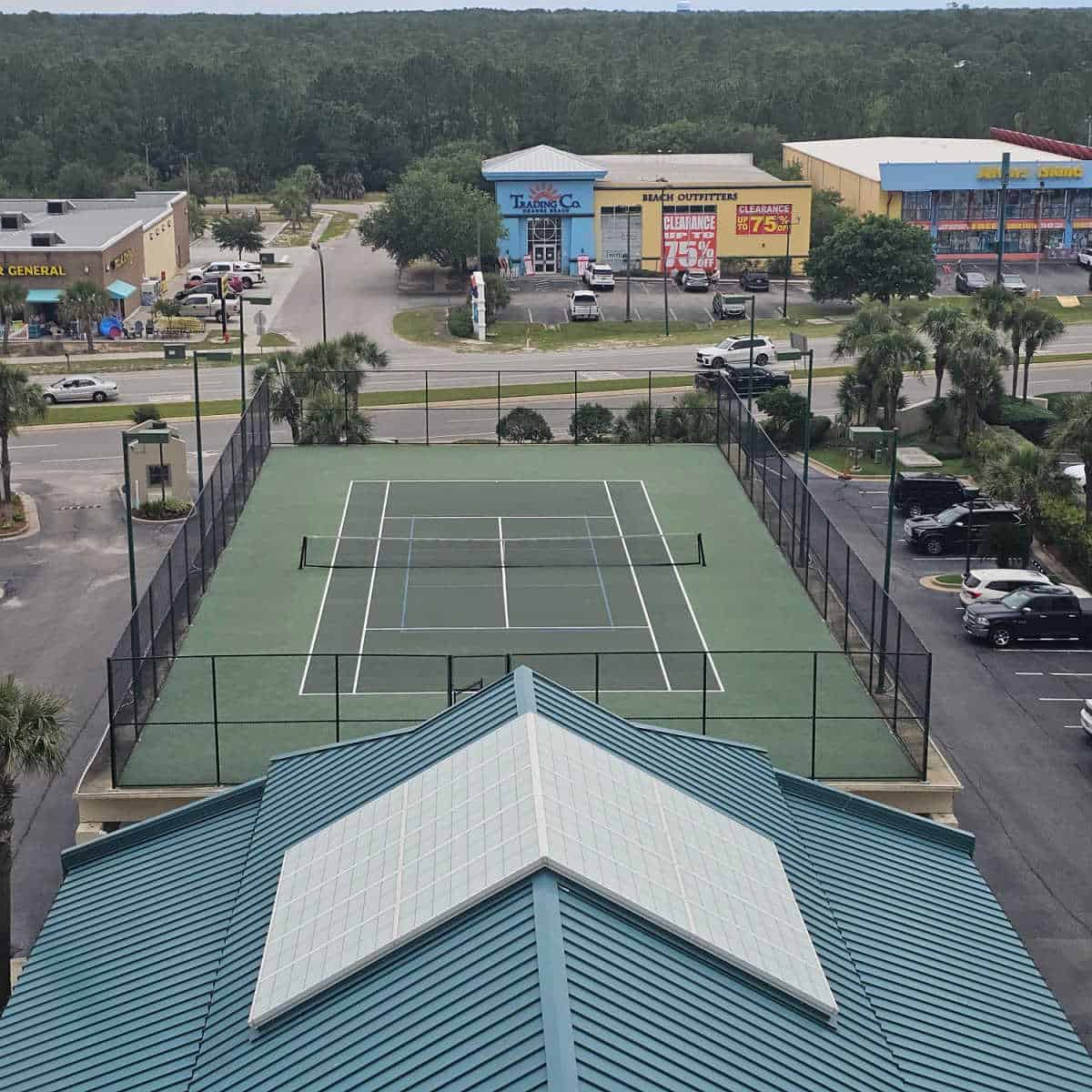 tennis court with fence around it, buildings across the street, cars in the parking lot