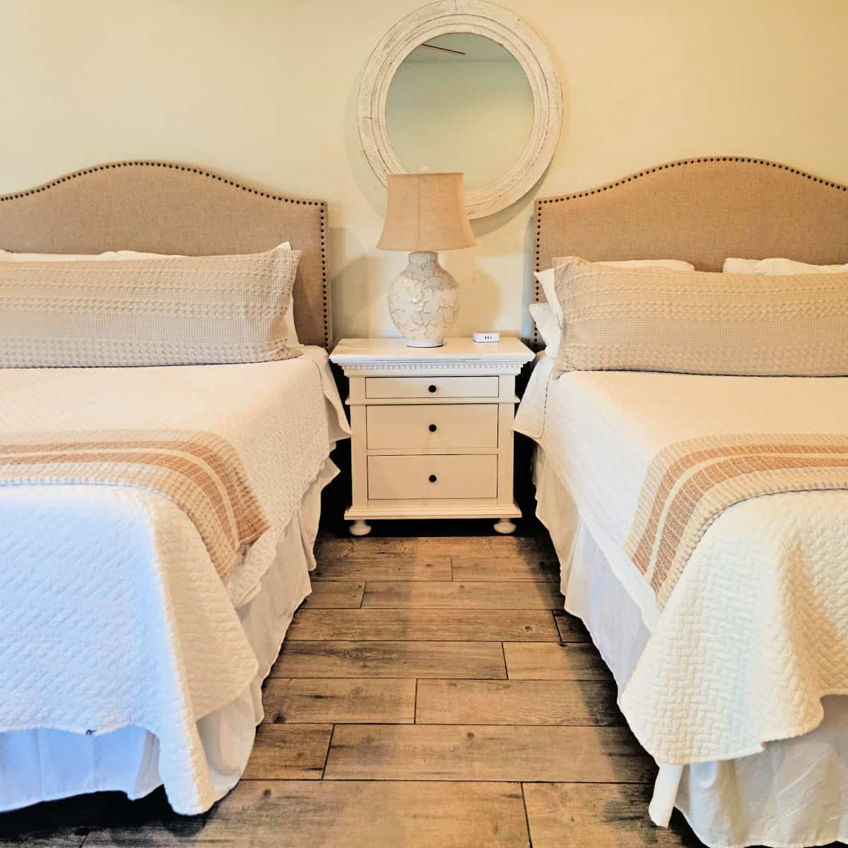 bedroom with two beds with white and tan linens, nightstand between the beds with a lamp and mirror on the wall
