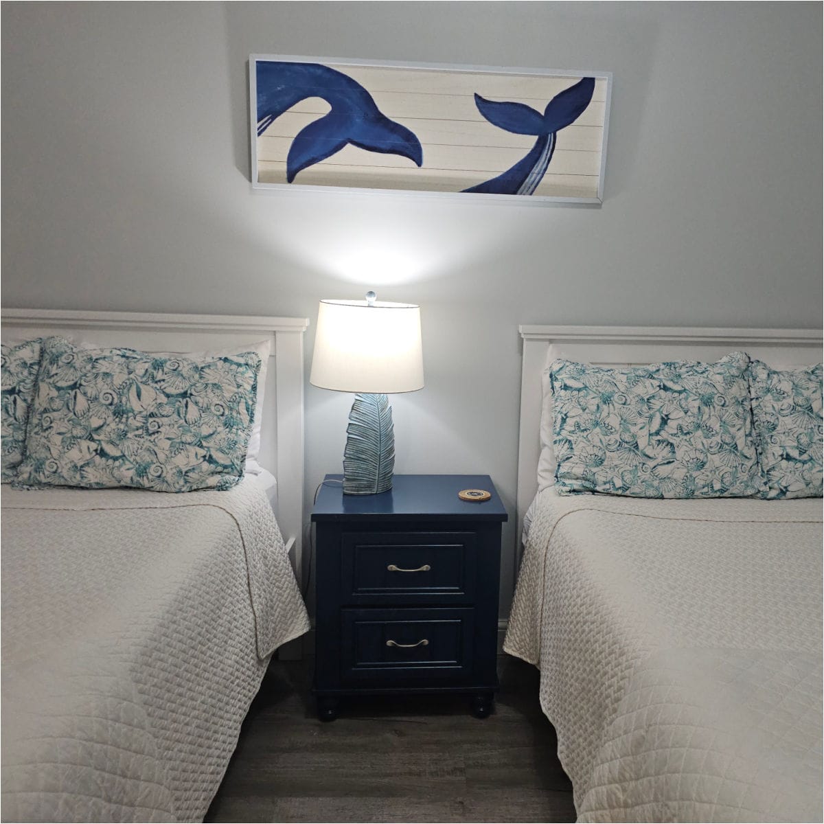two beds with white linens, blue shell pillowcases, blue nightstand with a lamp between the beds. Whales tale art above the lamp