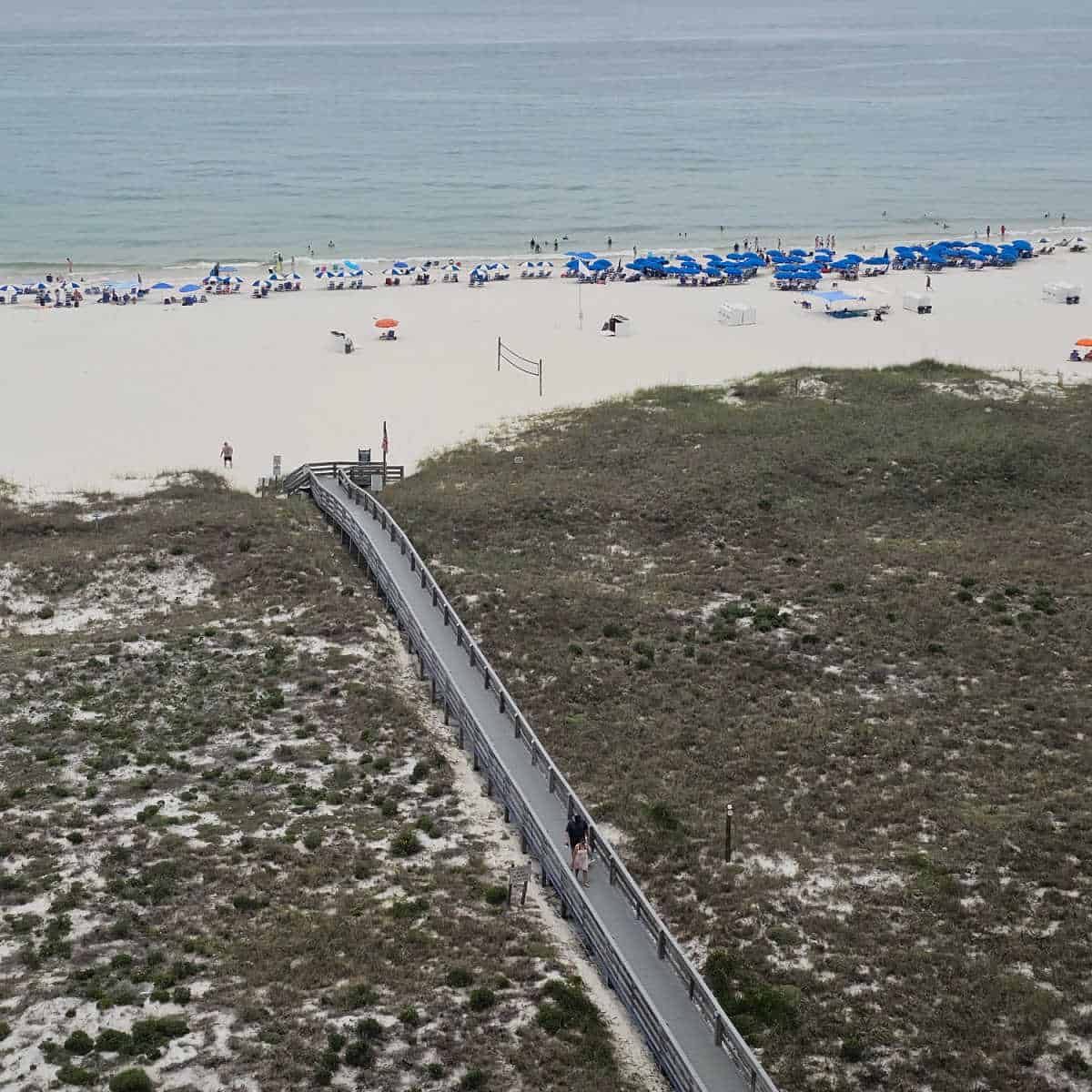 looking out over a boardwalk through sand dunes leading to the white sand beach and Gulf waters, blue and white umbrellas along the beach