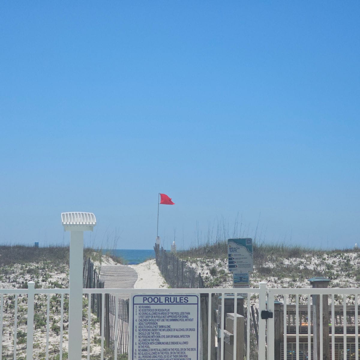 pool rules sign on a white fence, walkway through the sand to the beach with a red flag flying, gulf waters in the background