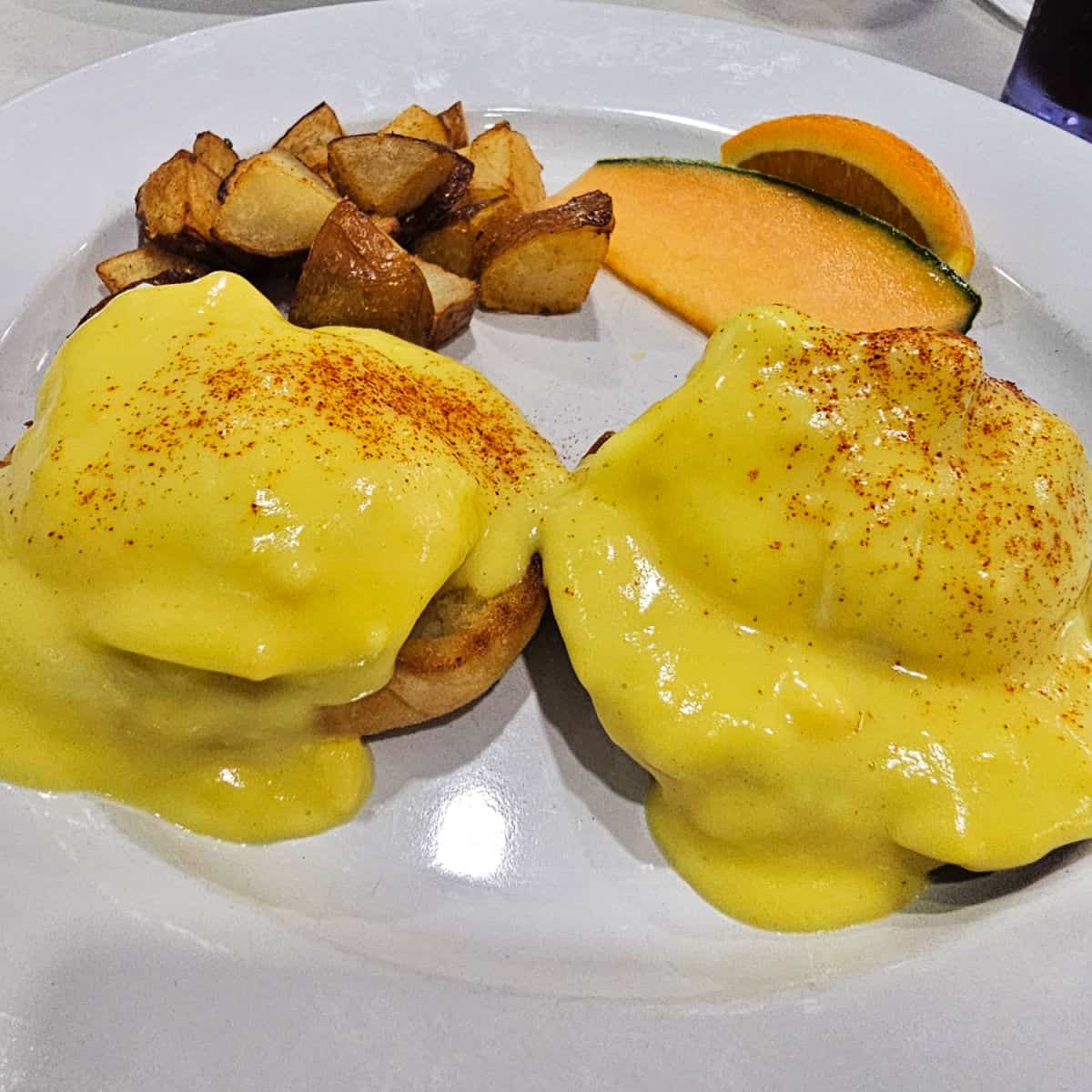 eggs benedict on a white plate with potatoes and a slice of cantoloupe