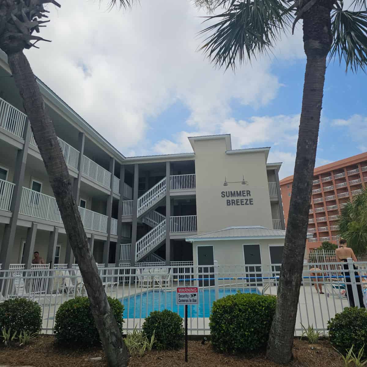 Summer breeze on the side of a 3 story building with white railing, pool on the ground level with a white fence around it and palm trees