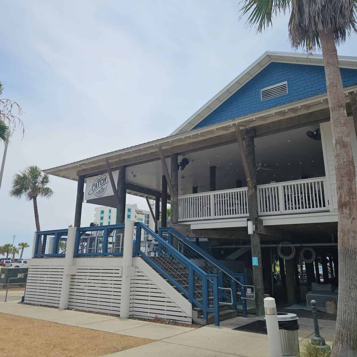 Exterior of The Catch restaurant with blue sides top, white railings and blue stair rails leading up to the restaurant, palm trees by the restaurant exterior
