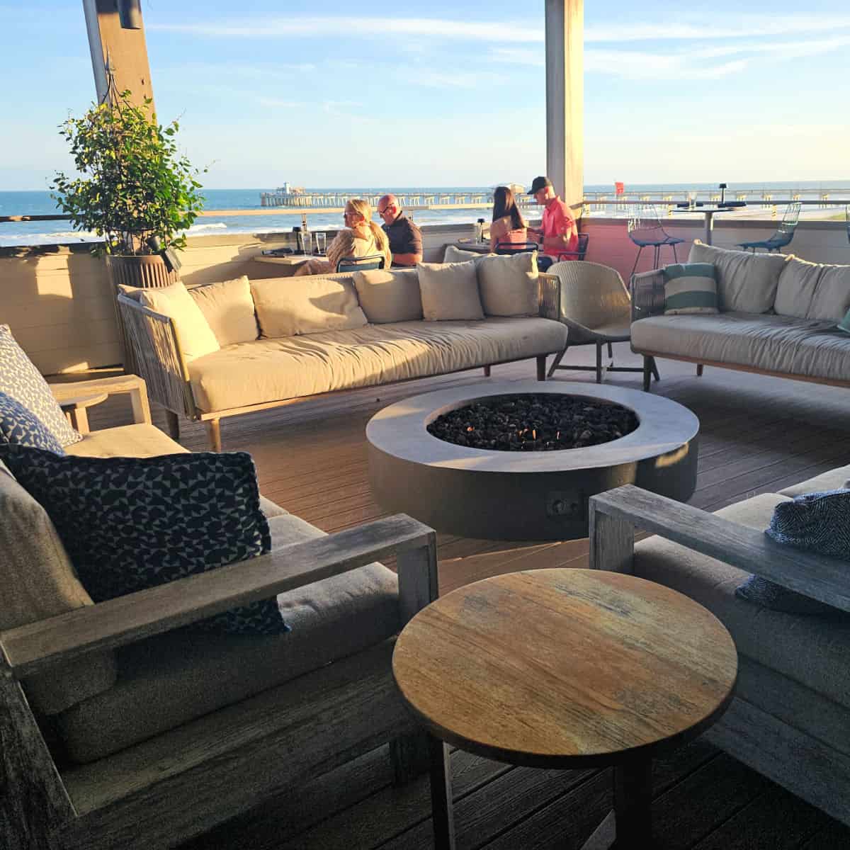 comfortable couches around a fire pit, people sitting at tables for two next to the balcony with a view of Gulf State Park pier