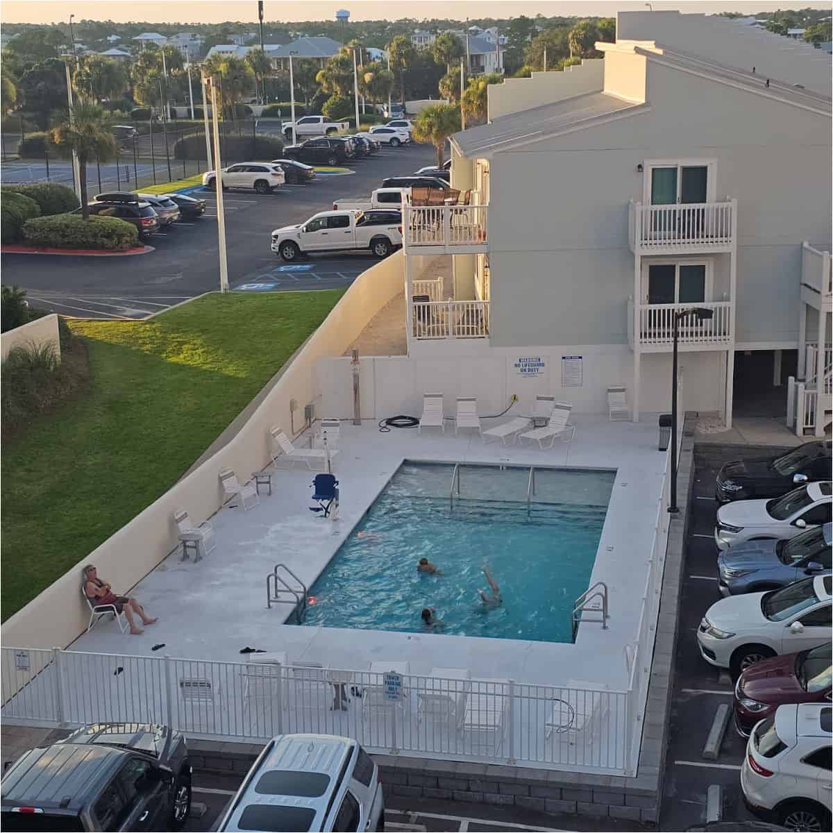 looking over to the pool with people playing in the water, lounge chairs around the deck, white building in the background with balconies, along with a parking lot and tennis court on the back left